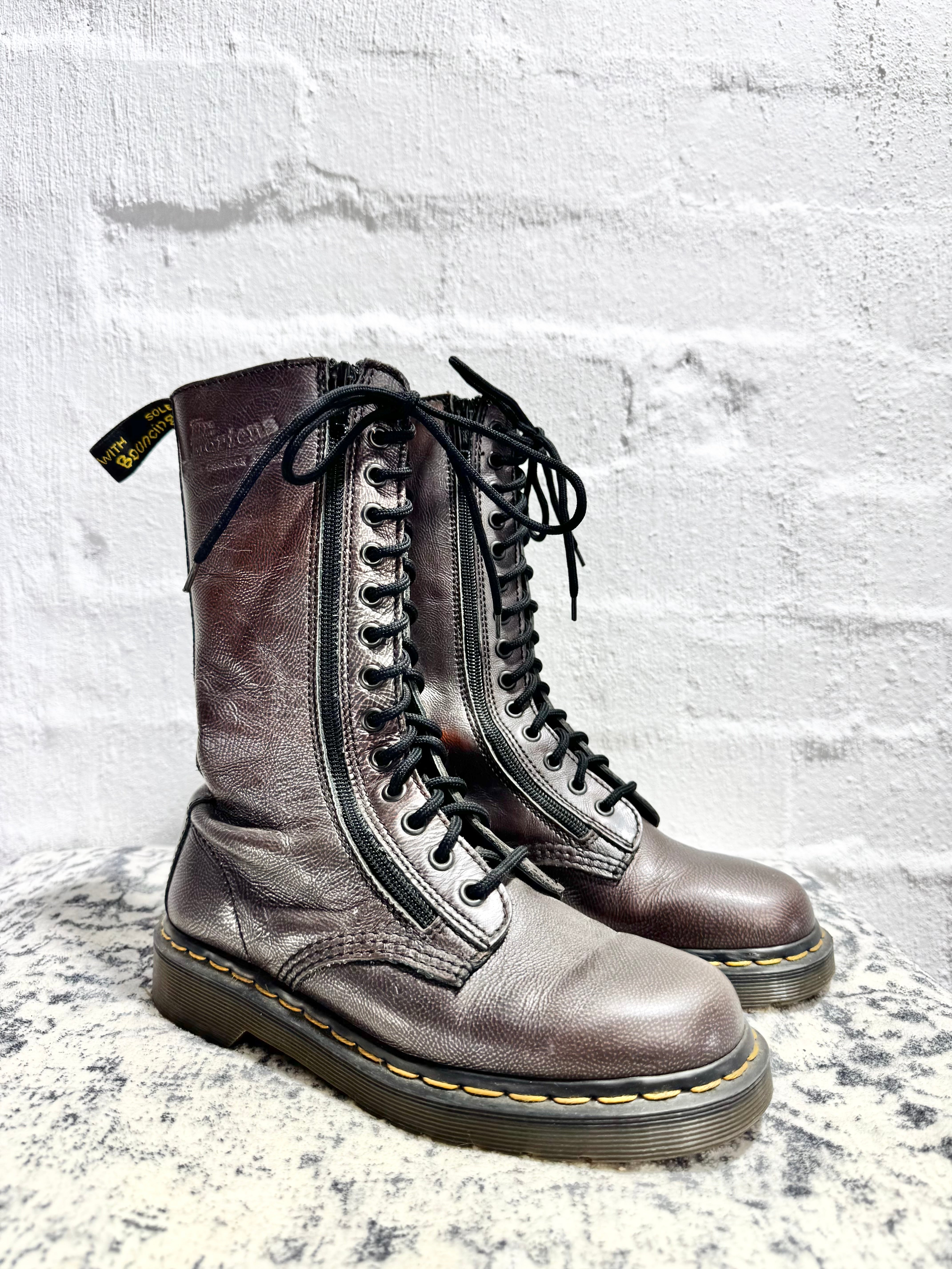 A pair of Dr. Martens 12-inch leather boots in a metallic bronze finish, displayed on a speckled stone surface against a white brick wall background.