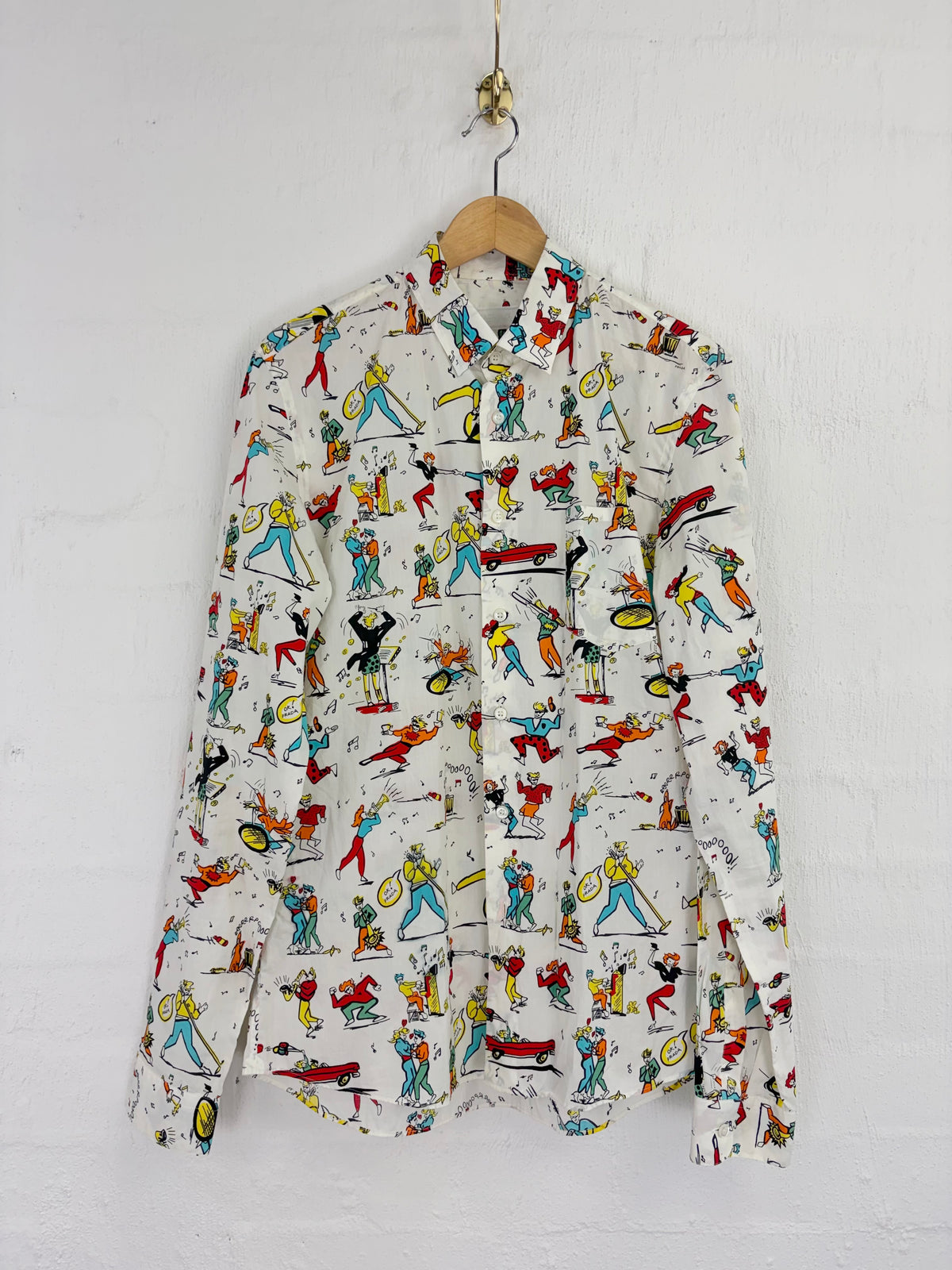 A long-sleeve button-up shirt with a white base and a colorful cartoon print of figures playing musical instruments, hanging on a wooden hanger against a white wall.
