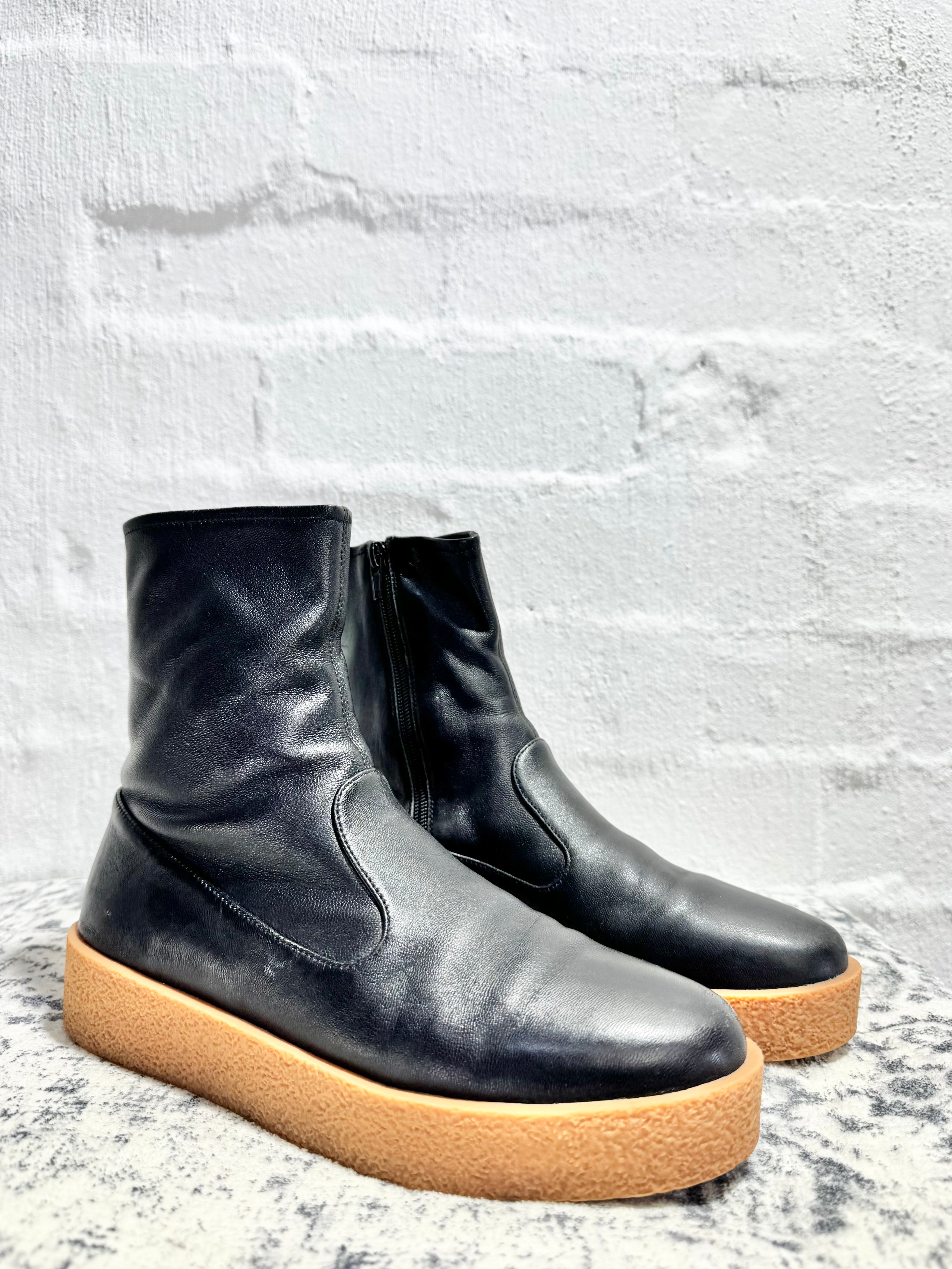 A pair of black leather ankle boots with chunky tan platform soles, displayed on a textured gray surface against a white brick wall background.