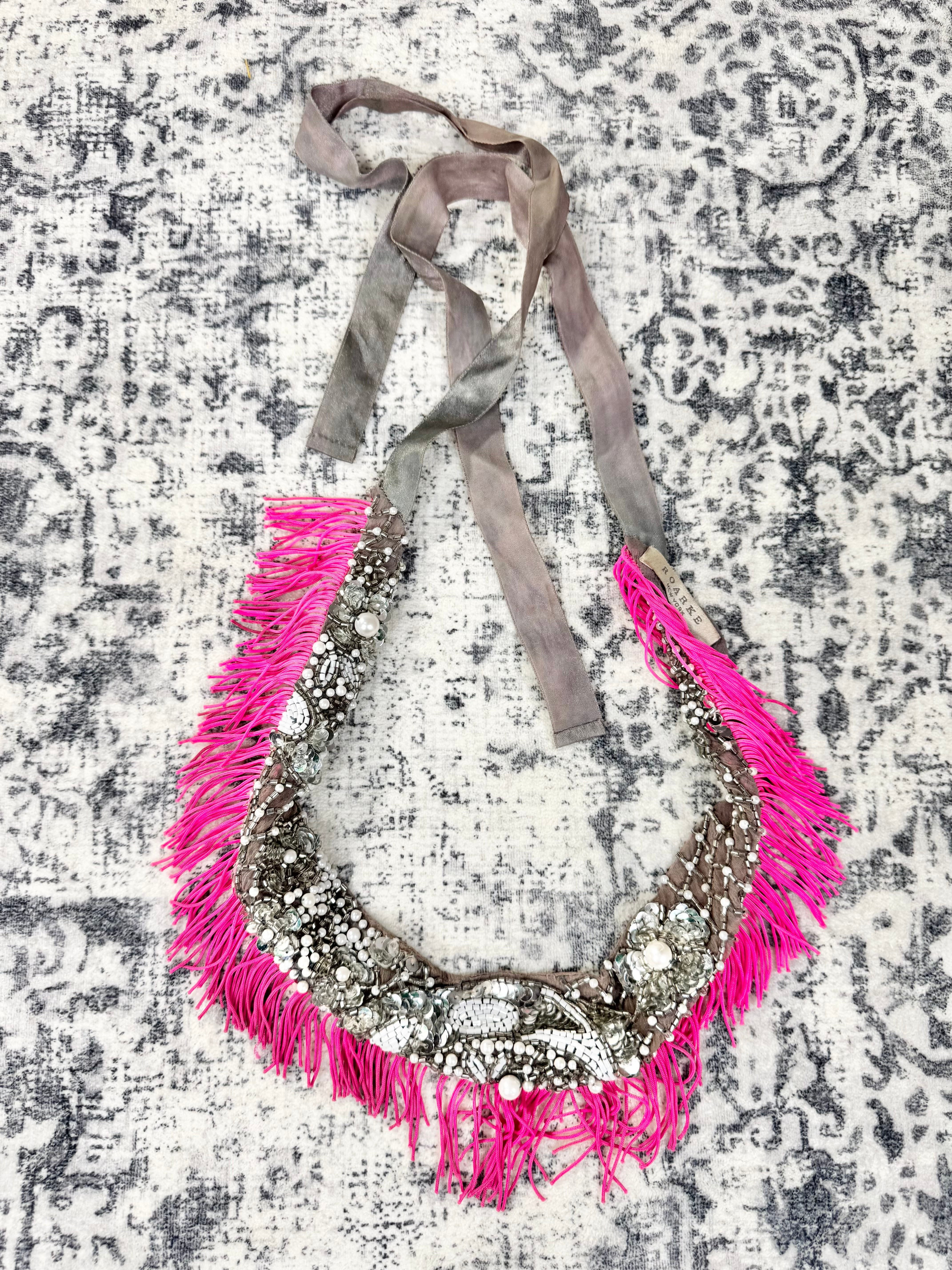 A bohemian-style halter necklace featuring a wide silver-toned metal collar covered in white beads, bright hot pink fringe, and matching pink fringe along the sides, with thin gray fabric ties at the top, laid flat on a distressed gray and white patterned rug.