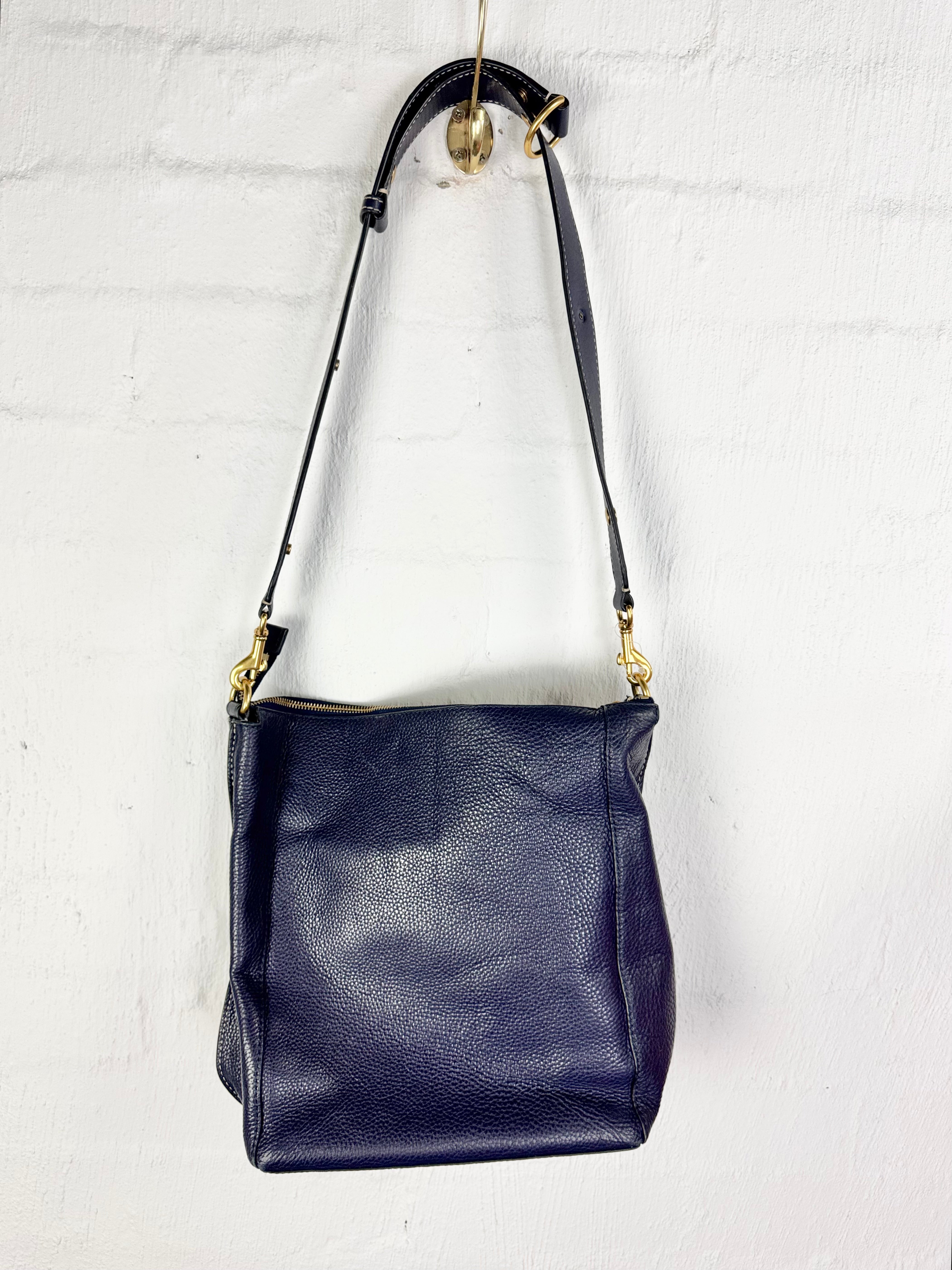 A navy blue pebbled leather shoulder bag hanging from a gold hook against a white painted brick wall. The bag features a wide body, a thin adjustable shoulder strap with gold-tone hardware, and a top zipper closure.