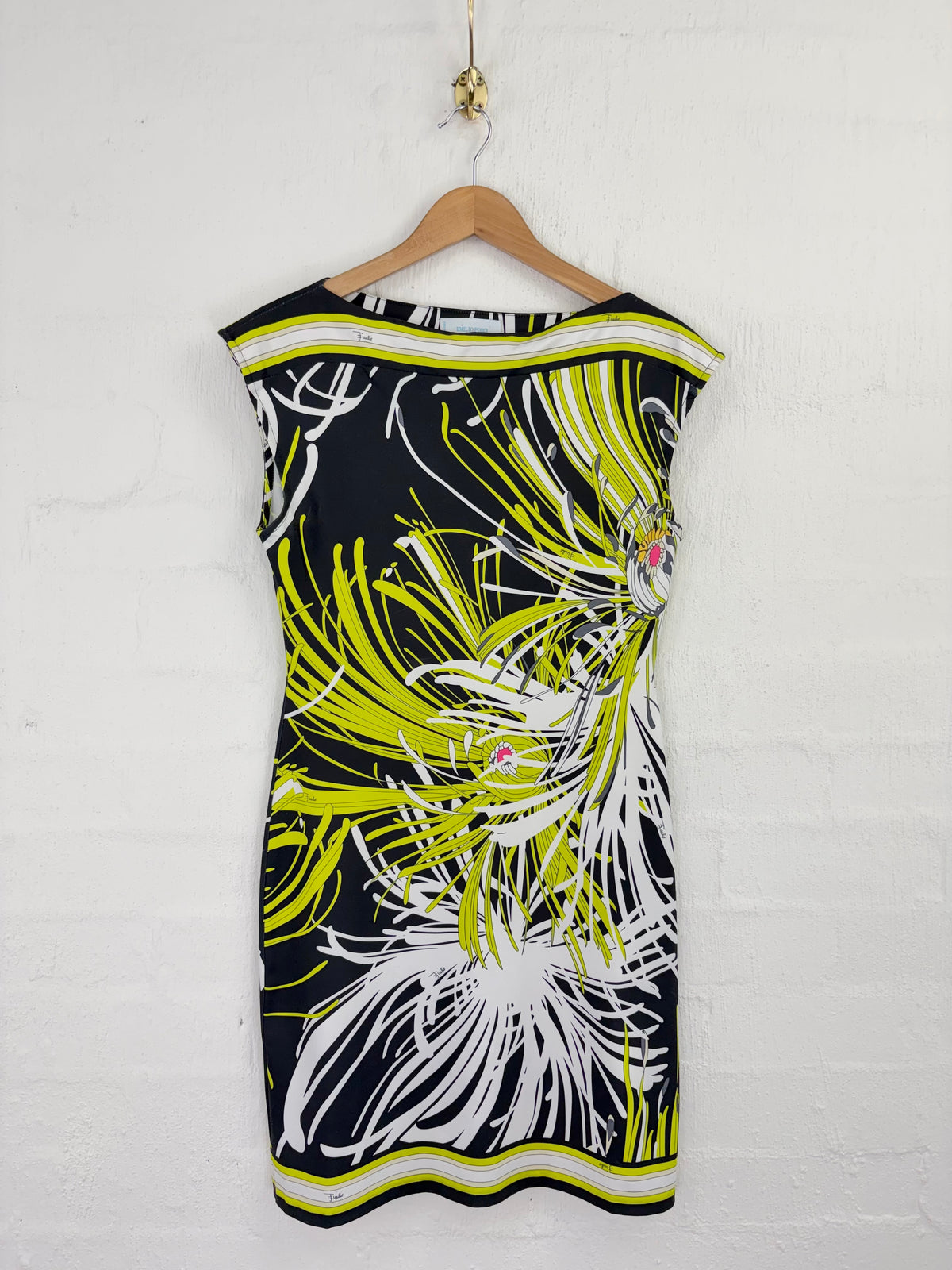 A sleeveless bodycon dress with a bold abstract floral print in lime green, white, and black, hanging on a wooden hanger against a white painted brick wall.