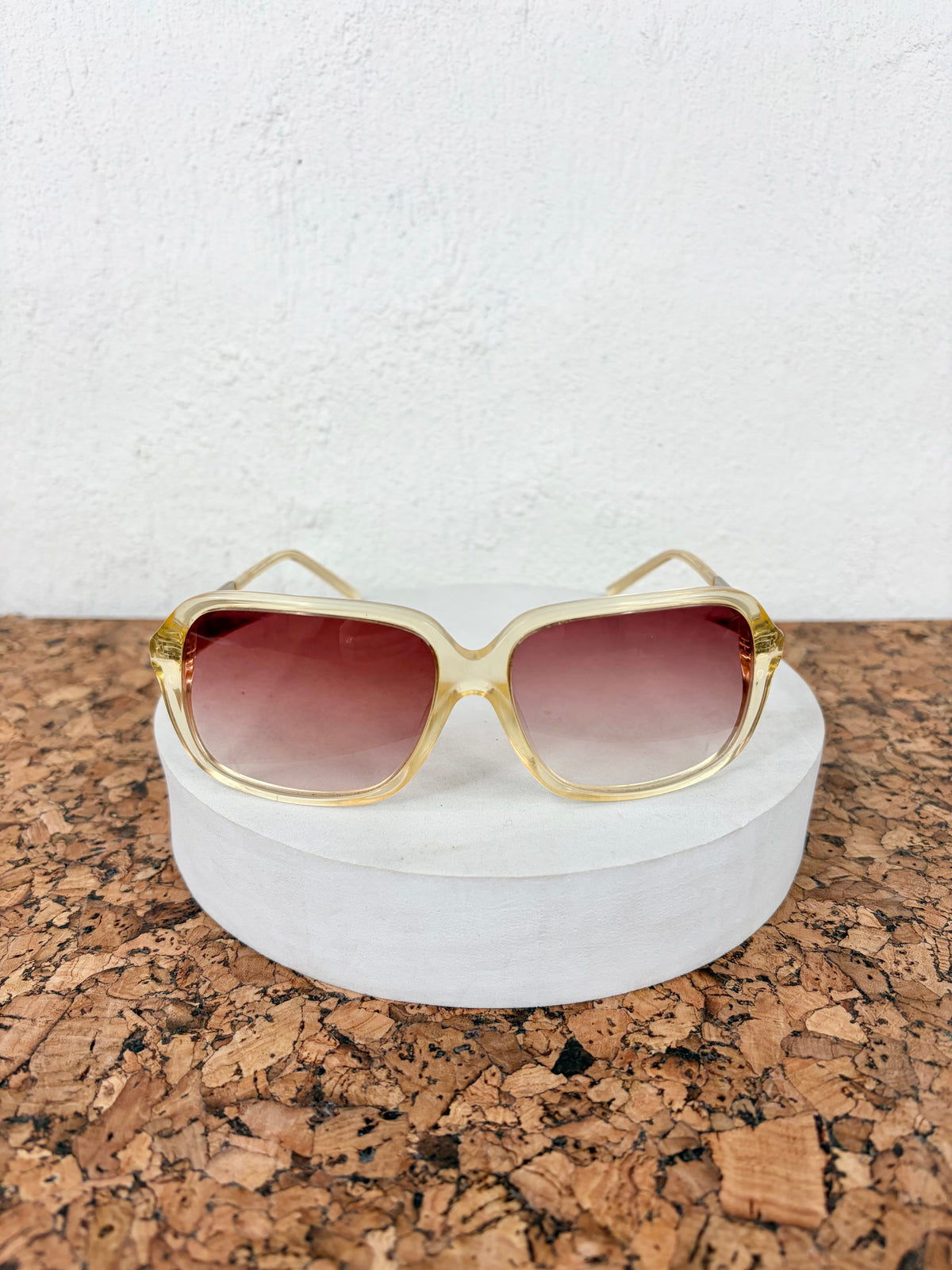 Oversized square sunglasses with a translucent light gold frame and gradient rose-tinted lenses, displayed on a round white pedestal over a cork surface against a white wall.