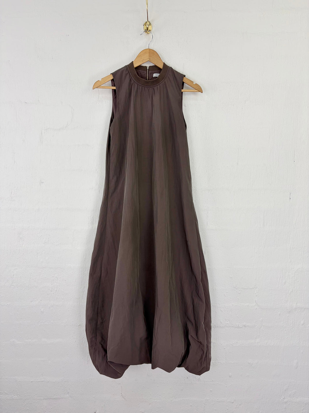 A sleeveless, high-neck maxi dress in a dark brownish-gray color, hanging on a wooden hanger against a white painted brick wall.