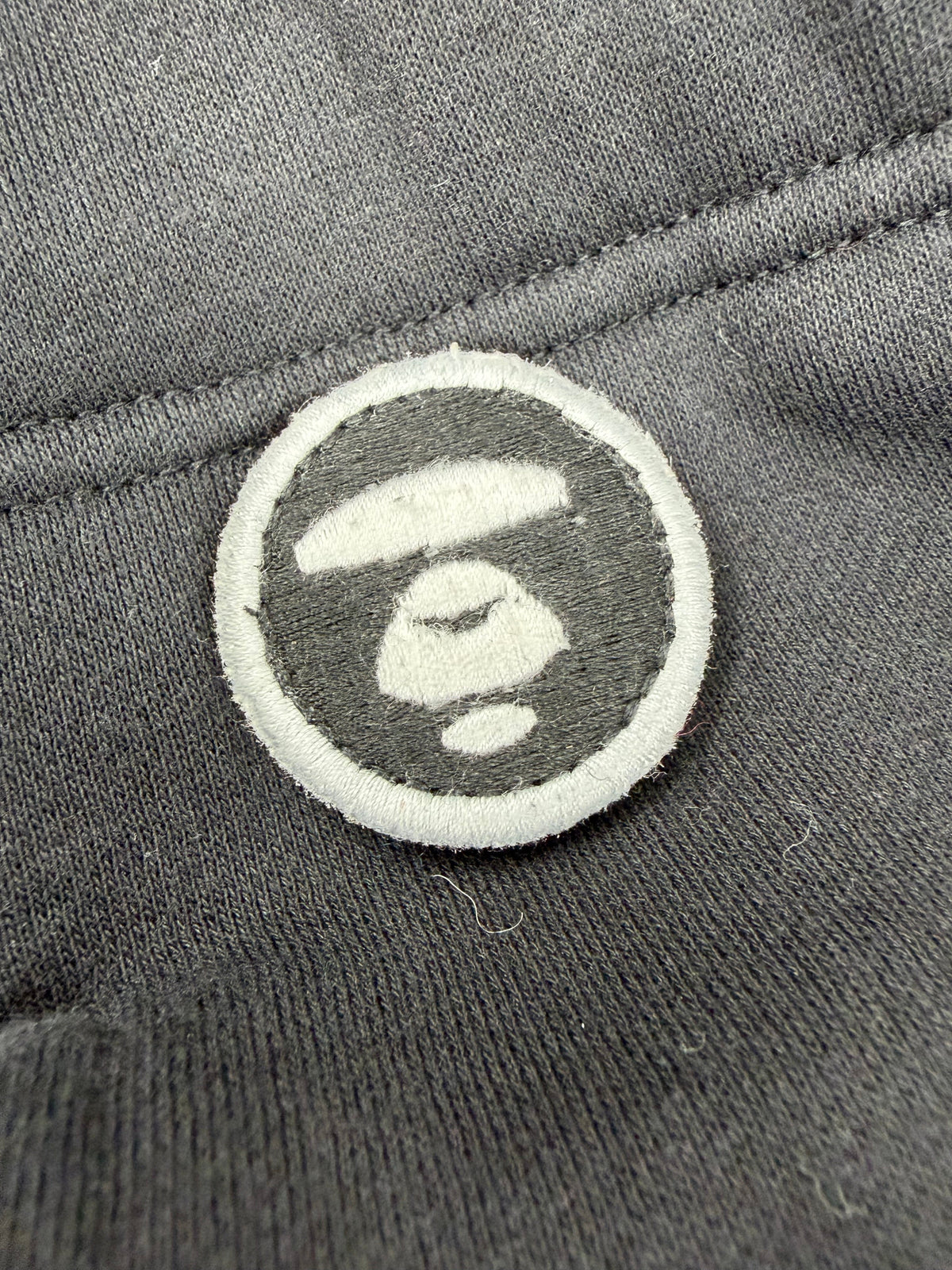 Close-up of a circular embroidered patch featuring a white stylized '10' logo on a dark gray background, attached to dark gray textured fabric.