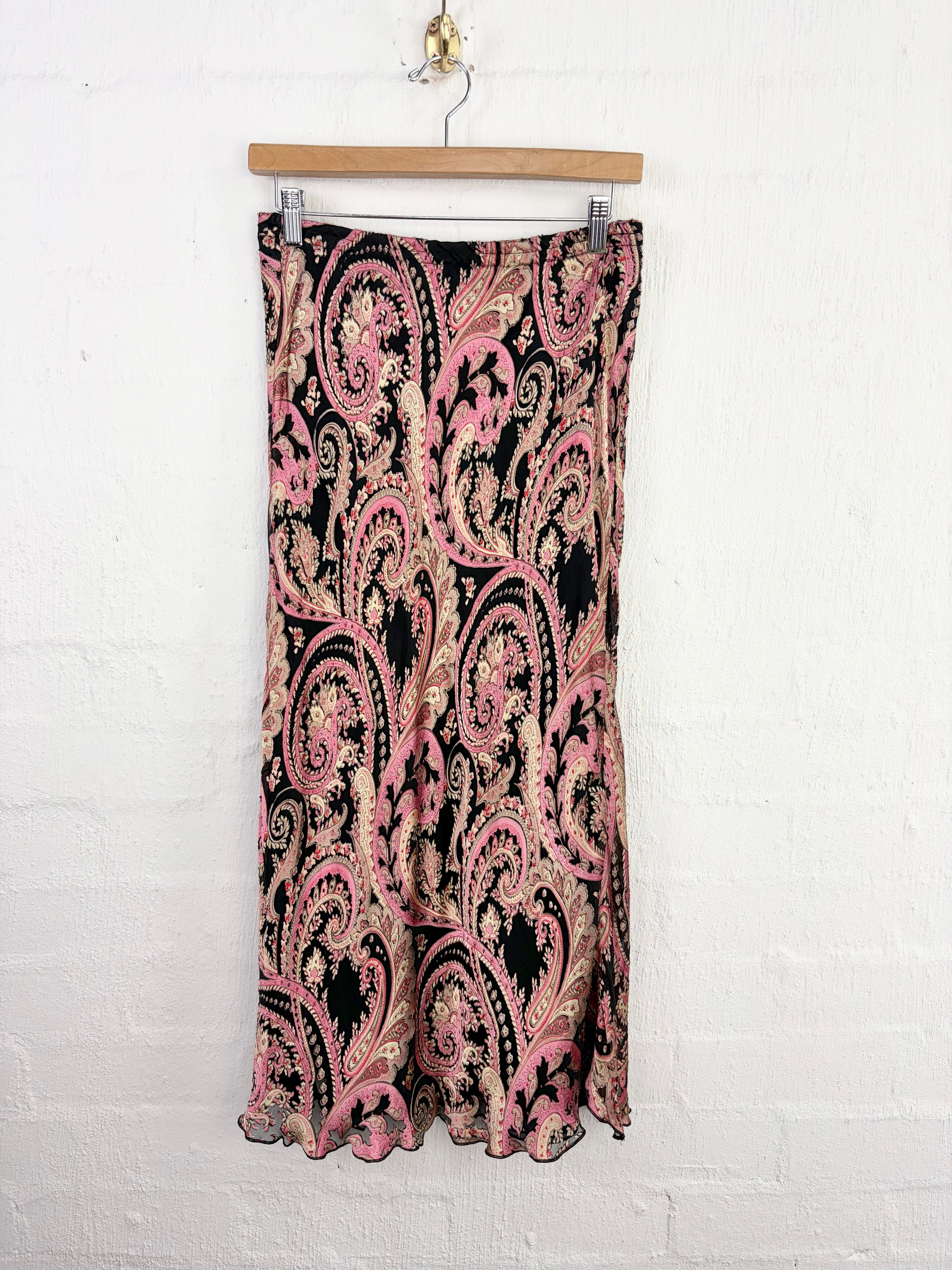 A black midi skirt with a pink and beige paisley pattern, hanging from a wooden hanger against a white brick wall.