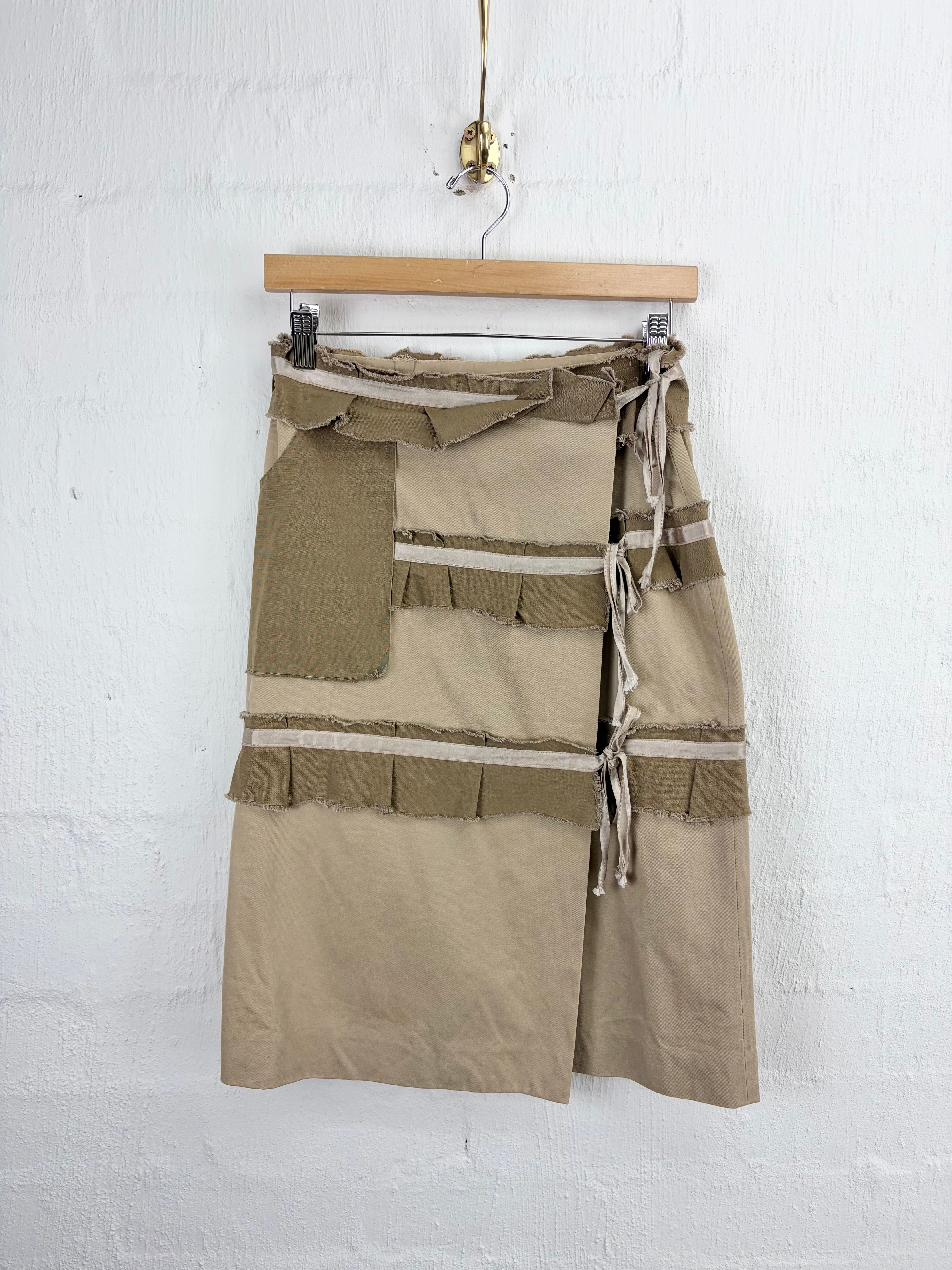 A beige and olive green patchwork skirt with ruffle details and side ties, hanging on a wooden hanger against a white textured wall.