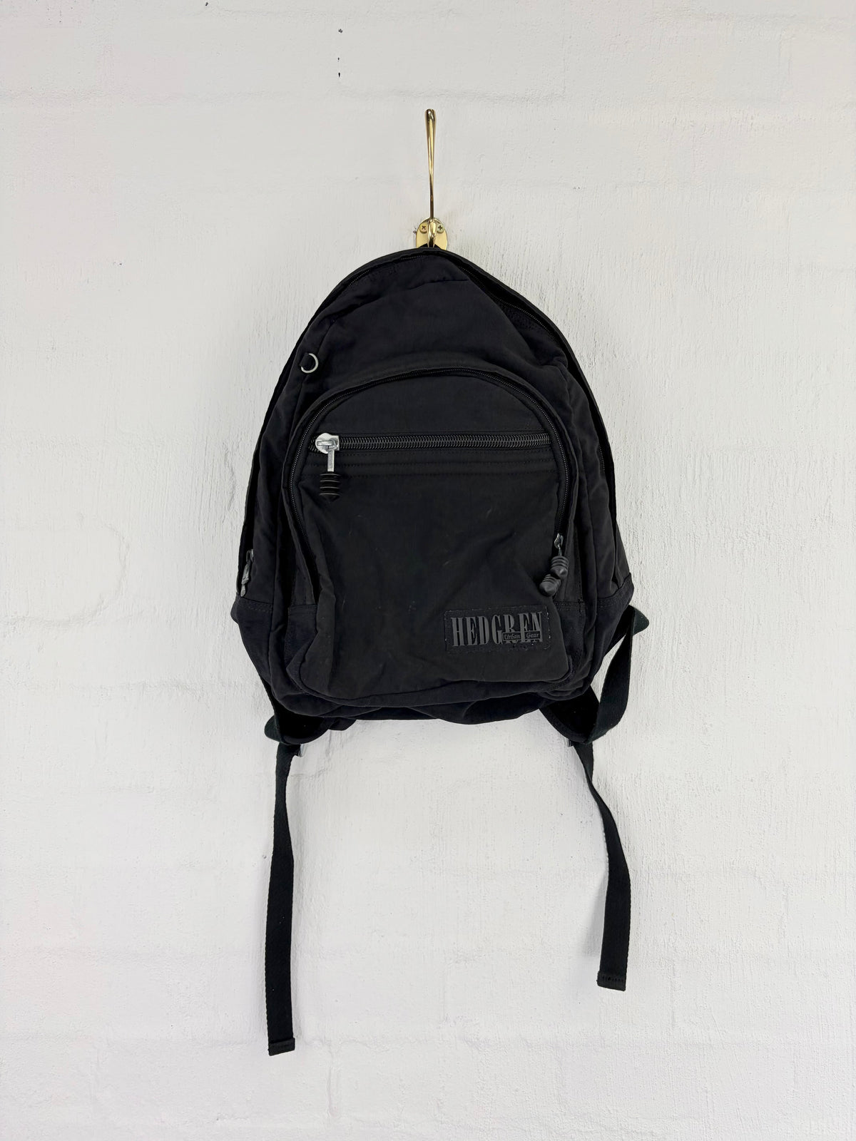 A black Hergex backpack hanging from a gold hook on a white brick wall. The backpack features a front zippered pocket with a silver zipper pull and a debossed black logo on the lower right of the pocket.