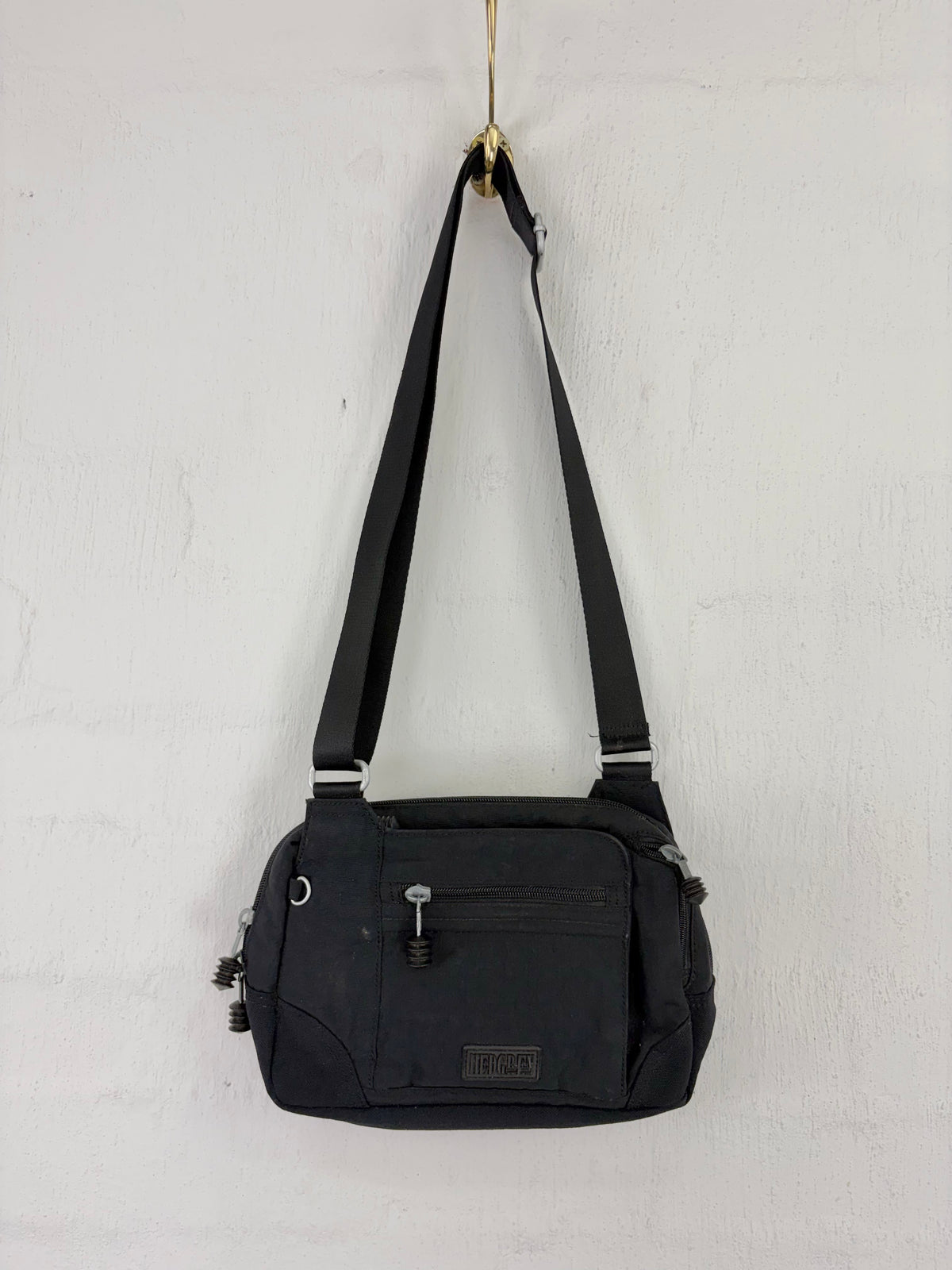 A black nylon shoulder bag with a long adjustable strap, hanging from a gold hook against a white textured wall. The bag features a front zippered pocket with a silver zipper pull and a small rectangular brand patch near the bottom.