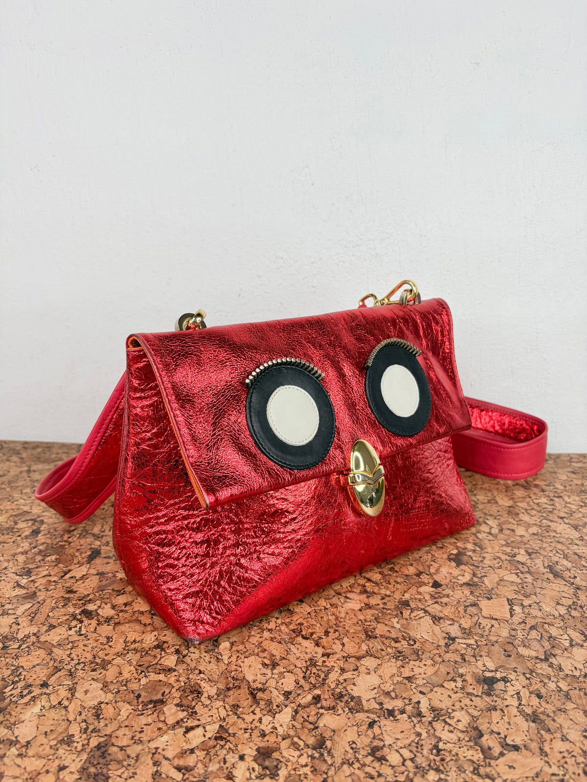 A metallic red handbag with a textured, crinkled finish, featuring large cartoon-style eyes and a gold clasp on the front flap. The bag has a matching red shoulder strap and is displayed on a cork surface against a plain white wall.