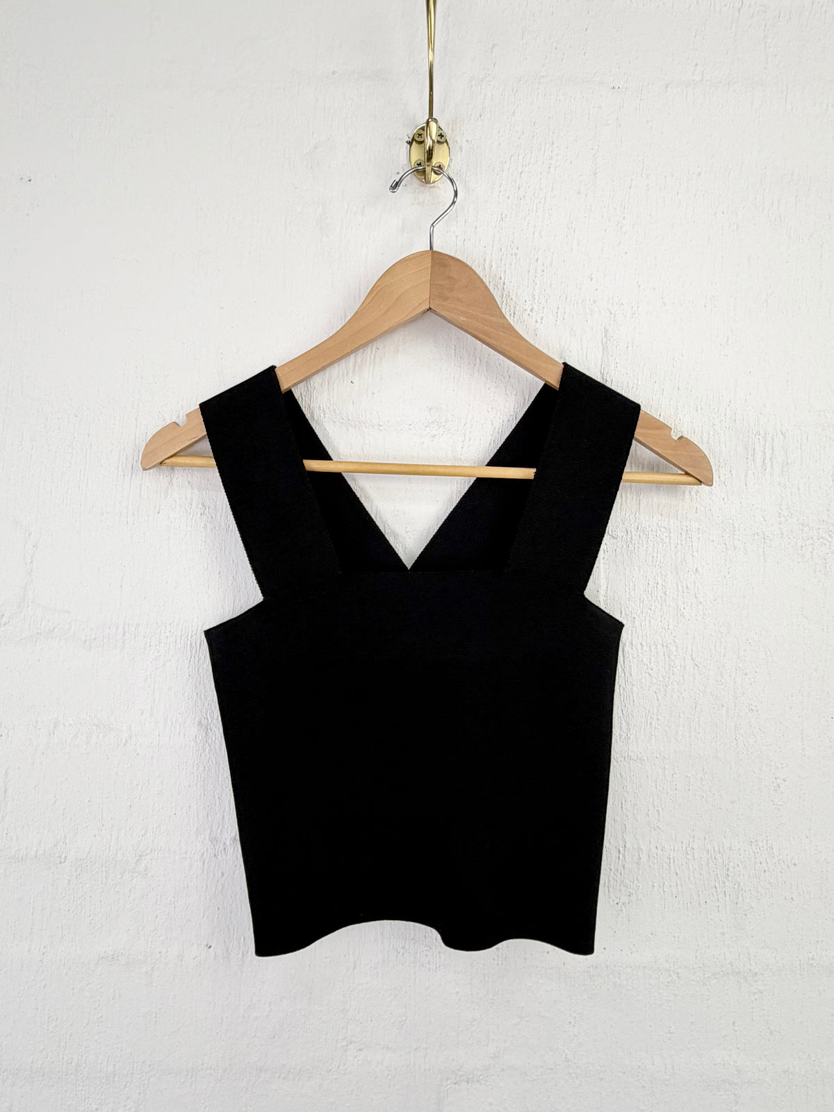 A black sleeveless knit top with wide straps and a V-neckline, hanging on a wooden hanger against a white textured wall.