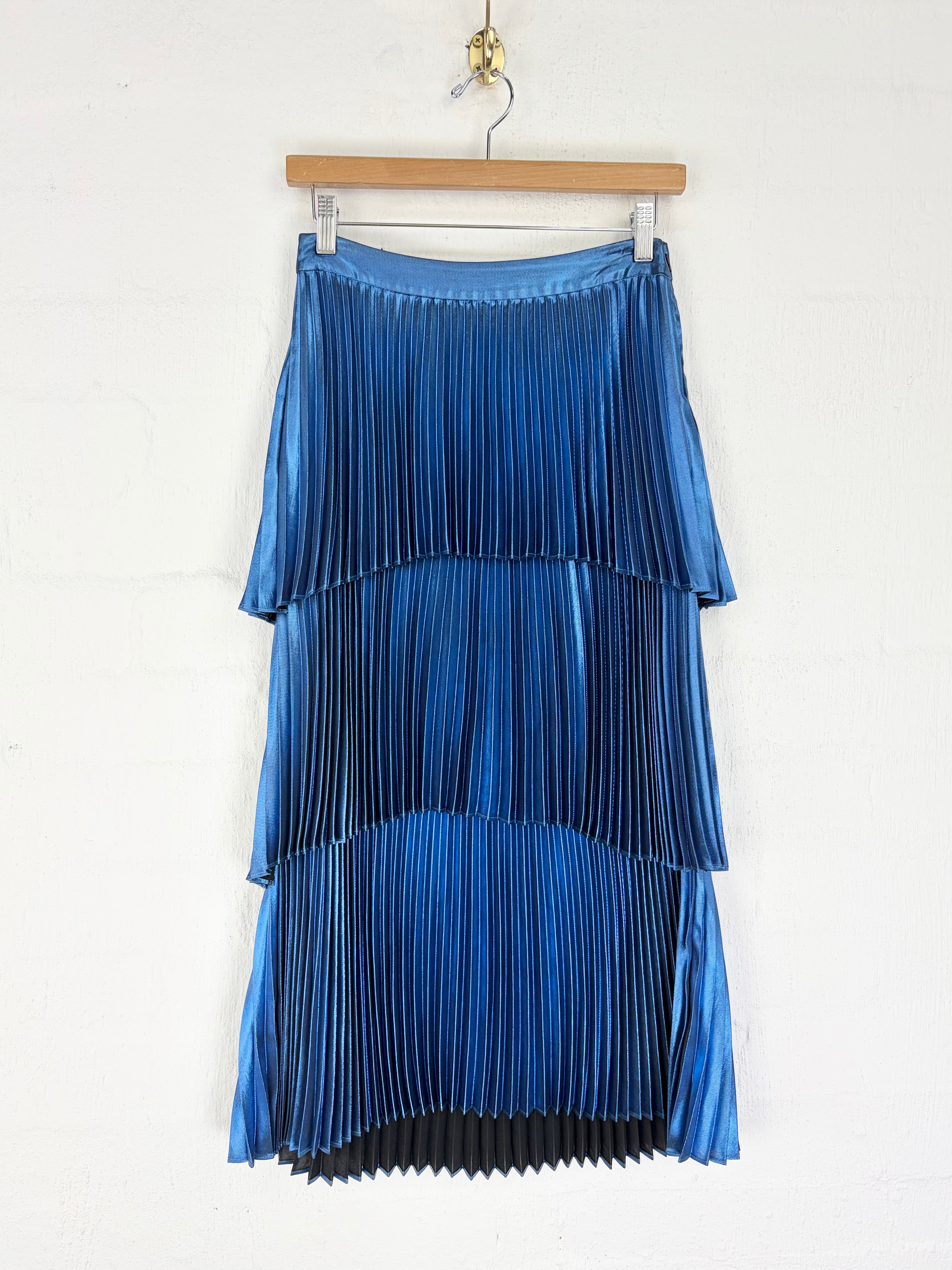 A tiered blue pleated midi skirt with a satin-like sheen, hanging from a wooden hanger against a white wall.