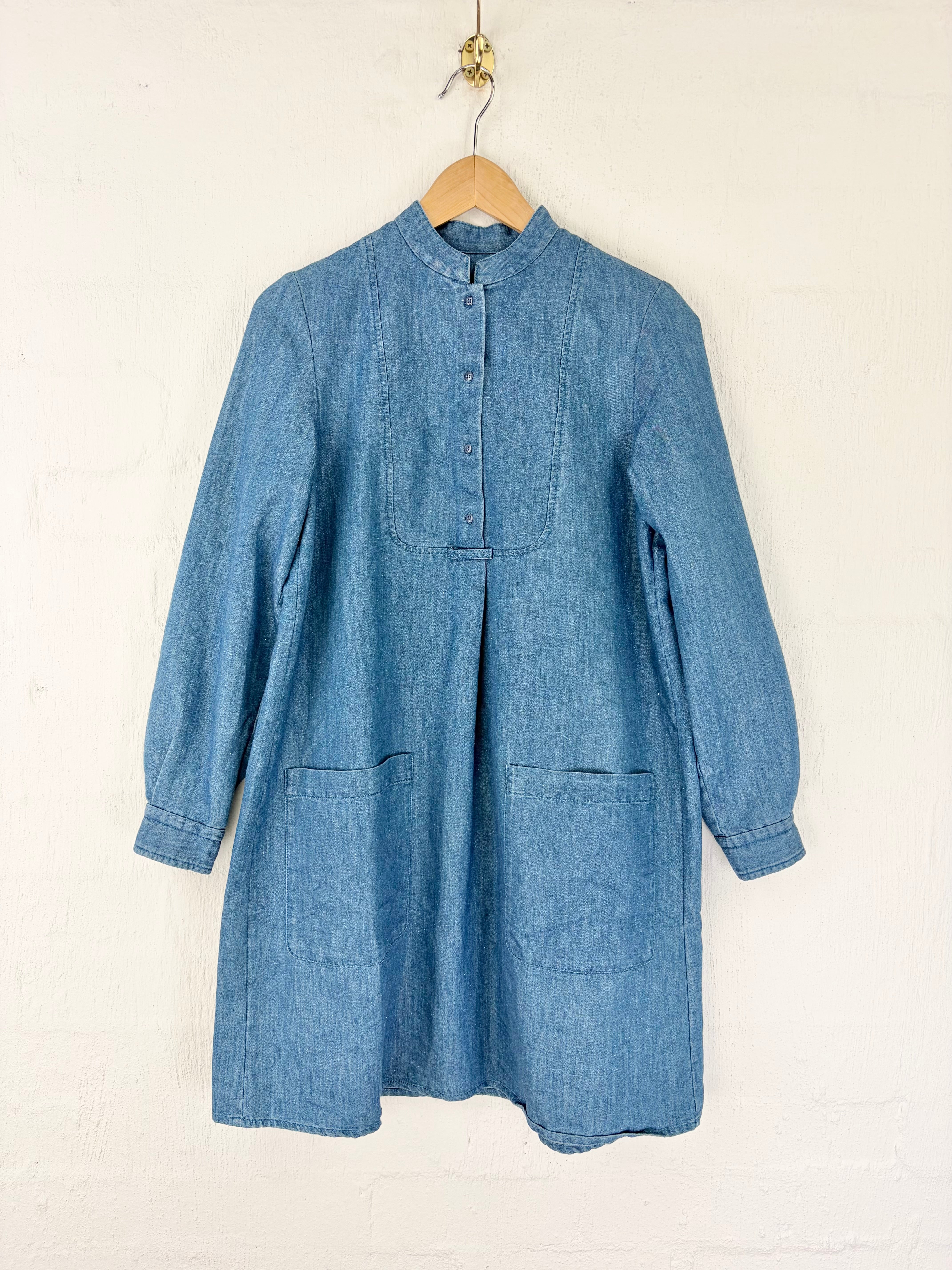 A blue denim long-sleeve tunic dress with a buttoned mandarin collar and front pockets, hanging on a wooden hanger against a white painted brick wall.