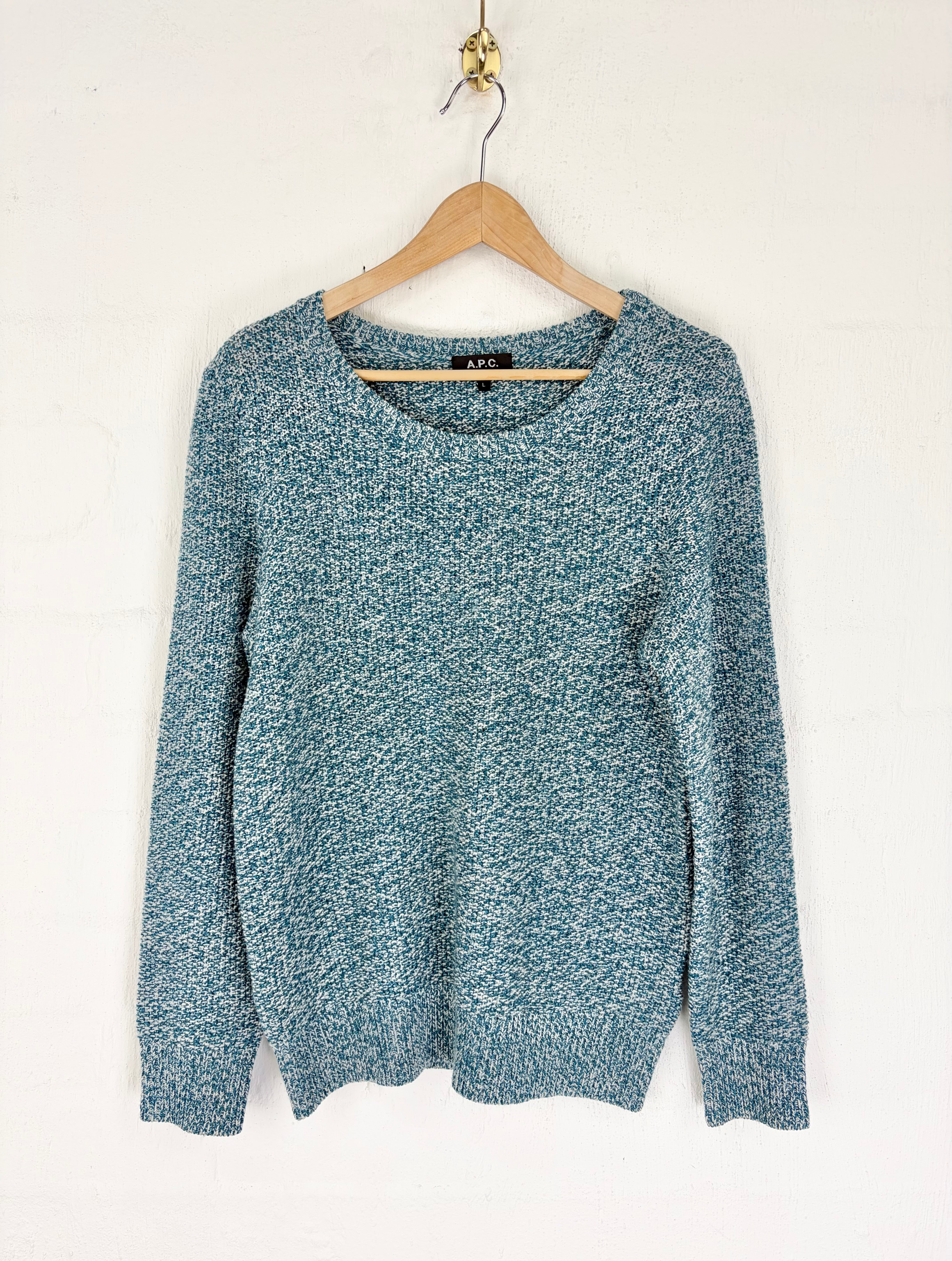 A long-sleeve knit sweater in a heathered teal blue and white marled pattern, hanging on a wooden hanger against a white wall.