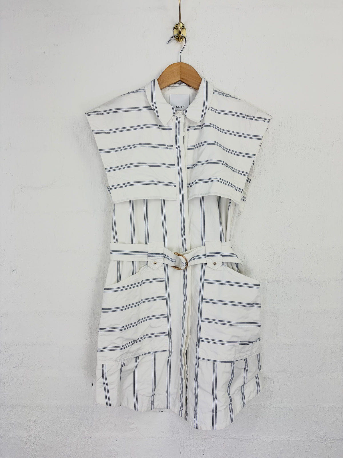 A sleeveless white linen-like top with thin blue vertical stripes, featuring a collar, front zipper, side cutouts, and a matching tie belt, hanging on a wooden hanger against a white wall.