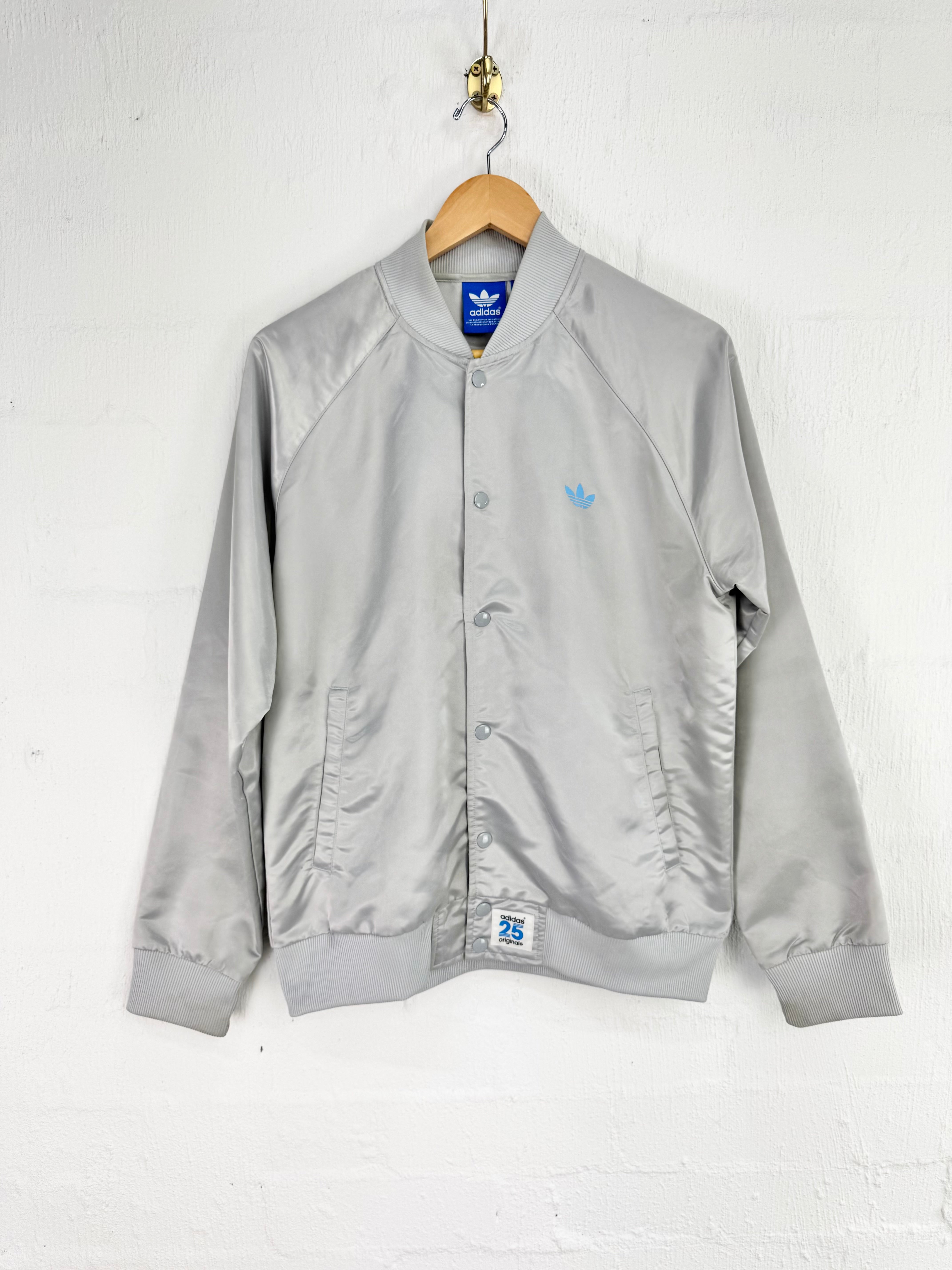 A light gray satin bomber jacket from Adidas, hanging on a wooden hanger against a white brick wall. The jacket features a light blue embroidered Adidas trefoil logo on the left chest and a blue brand label at the collar.