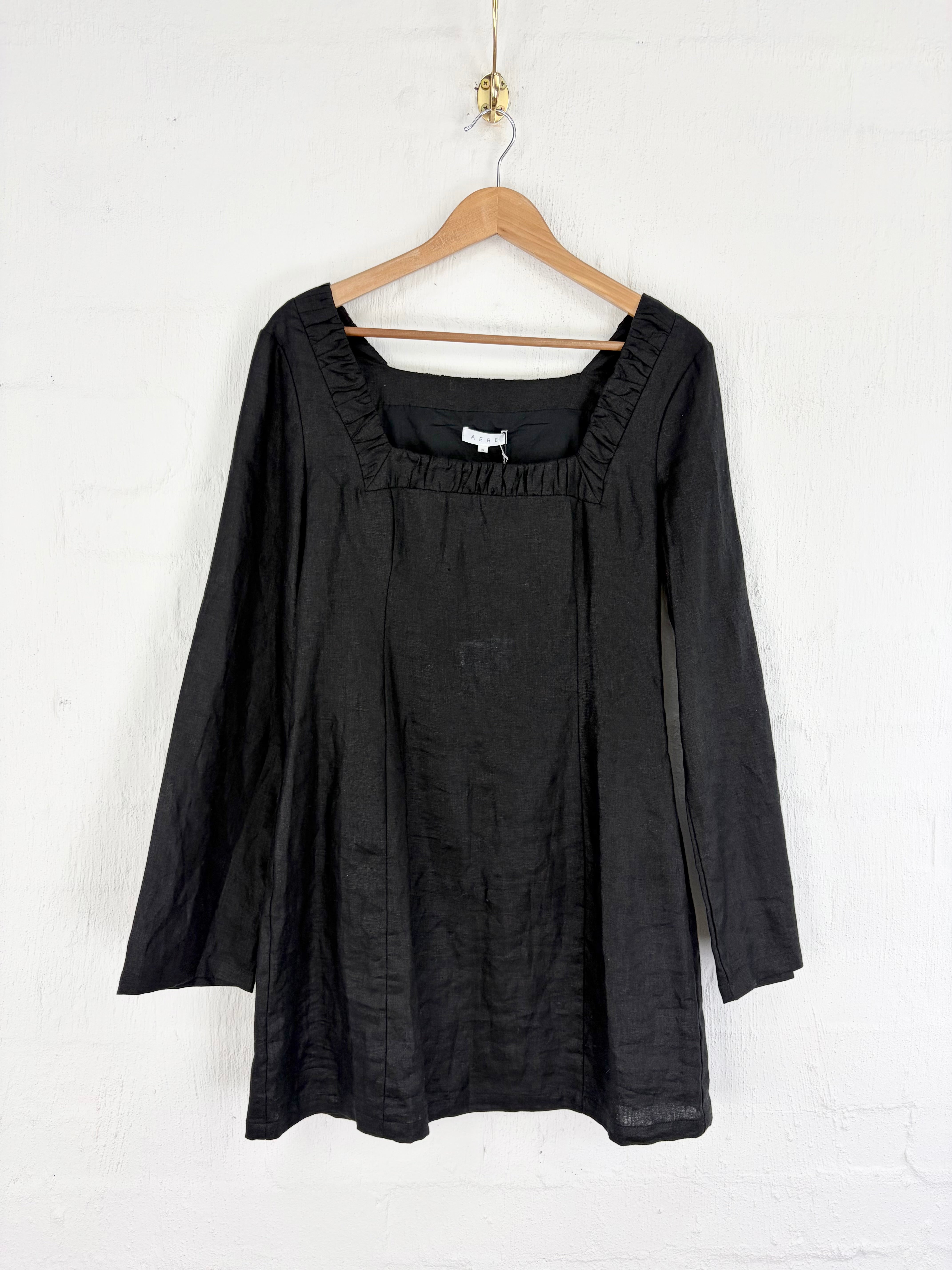 A black long-sleeve linen top with a square neckline and gathered ruffle trim, hanging on a wooden hanger against a white painted brick wall.