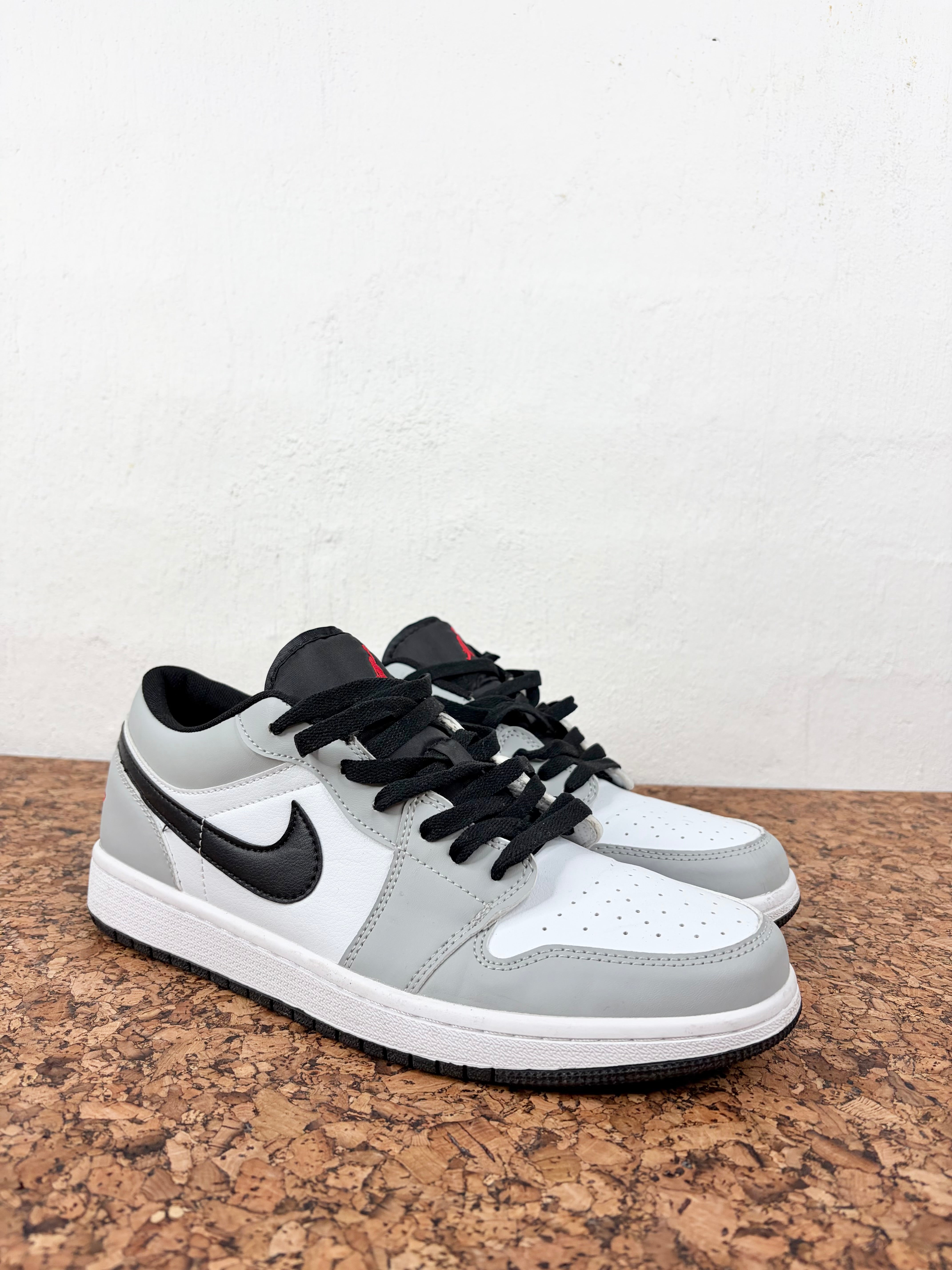 A pair of low-top Nike sneakers in light gray, white, and black, displayed on a cork mat against a white wall.