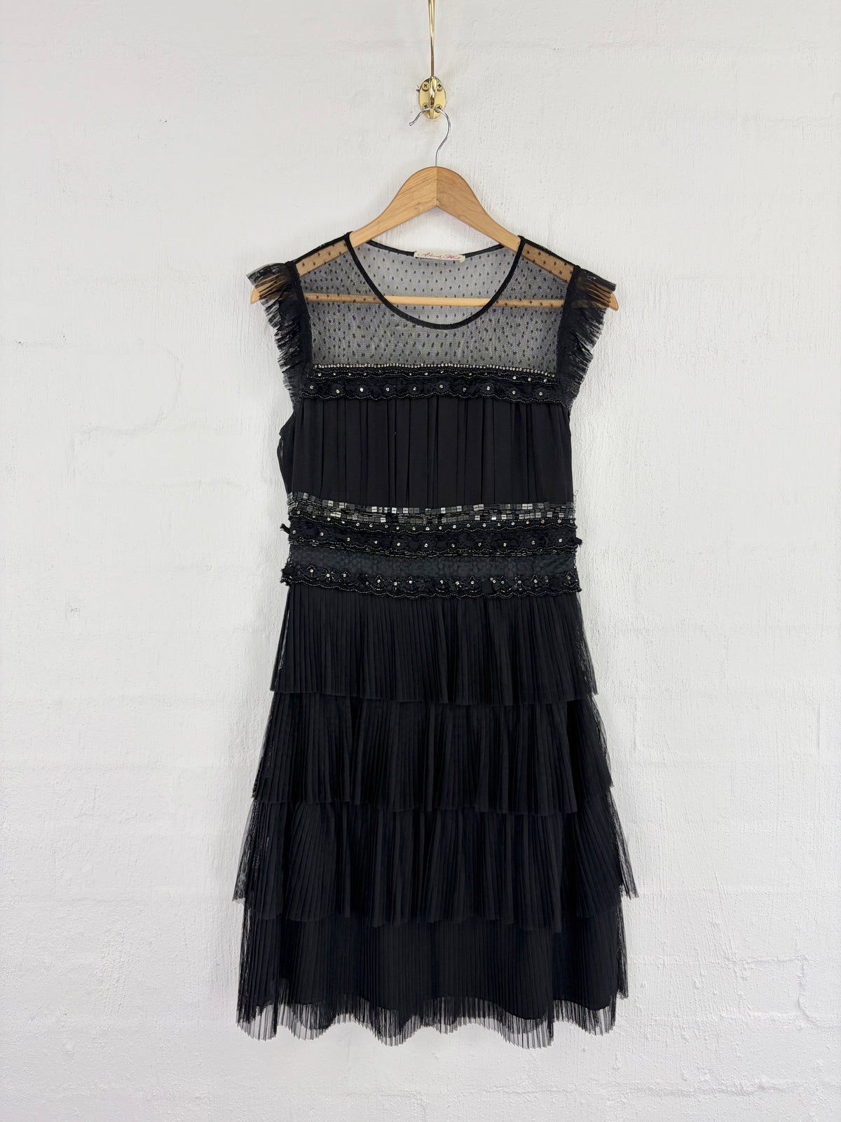A sleeveless black dress with a sheer mesh yoke, beaded horizontal detail at the waist, and a tiered tulle skirt, hanging on a wooden hanger against a white brick wall.