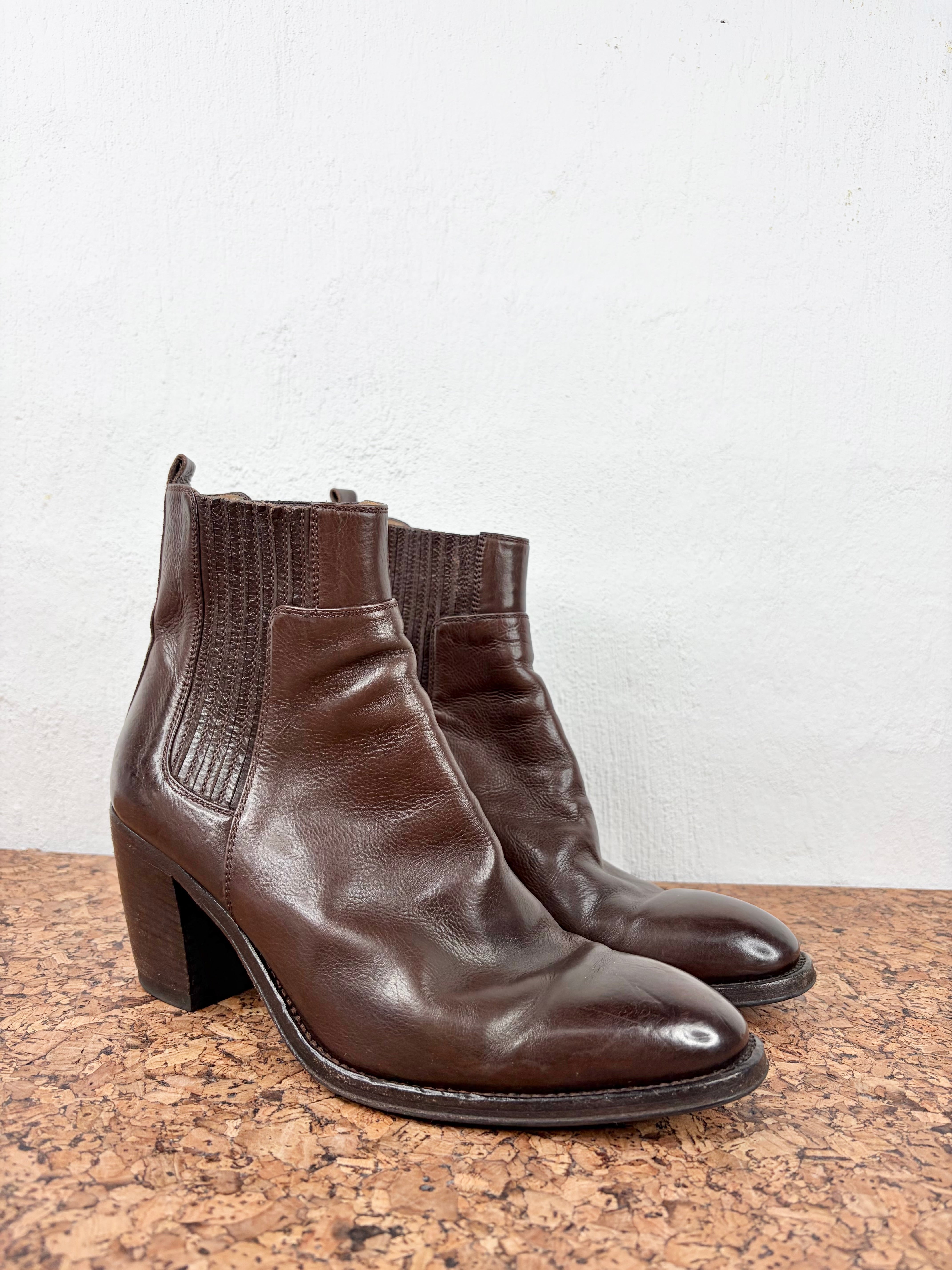 Pair of chocolate brown ankle boots on a corkboard surface against a white wall background