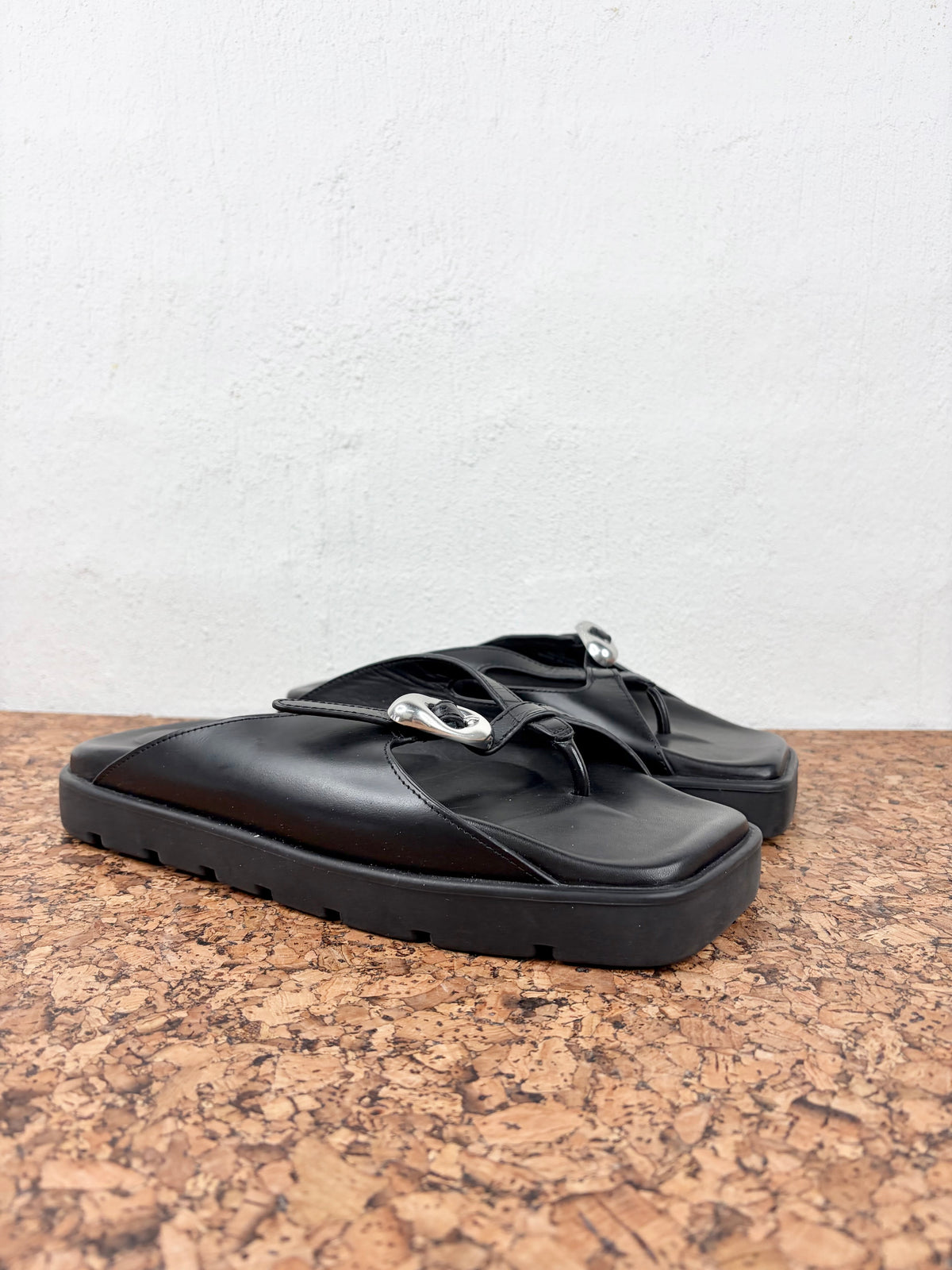 A pair of black leather slide sandals with chunky soles and silver-tone hardware, displayed on a cork surface against a white wall.