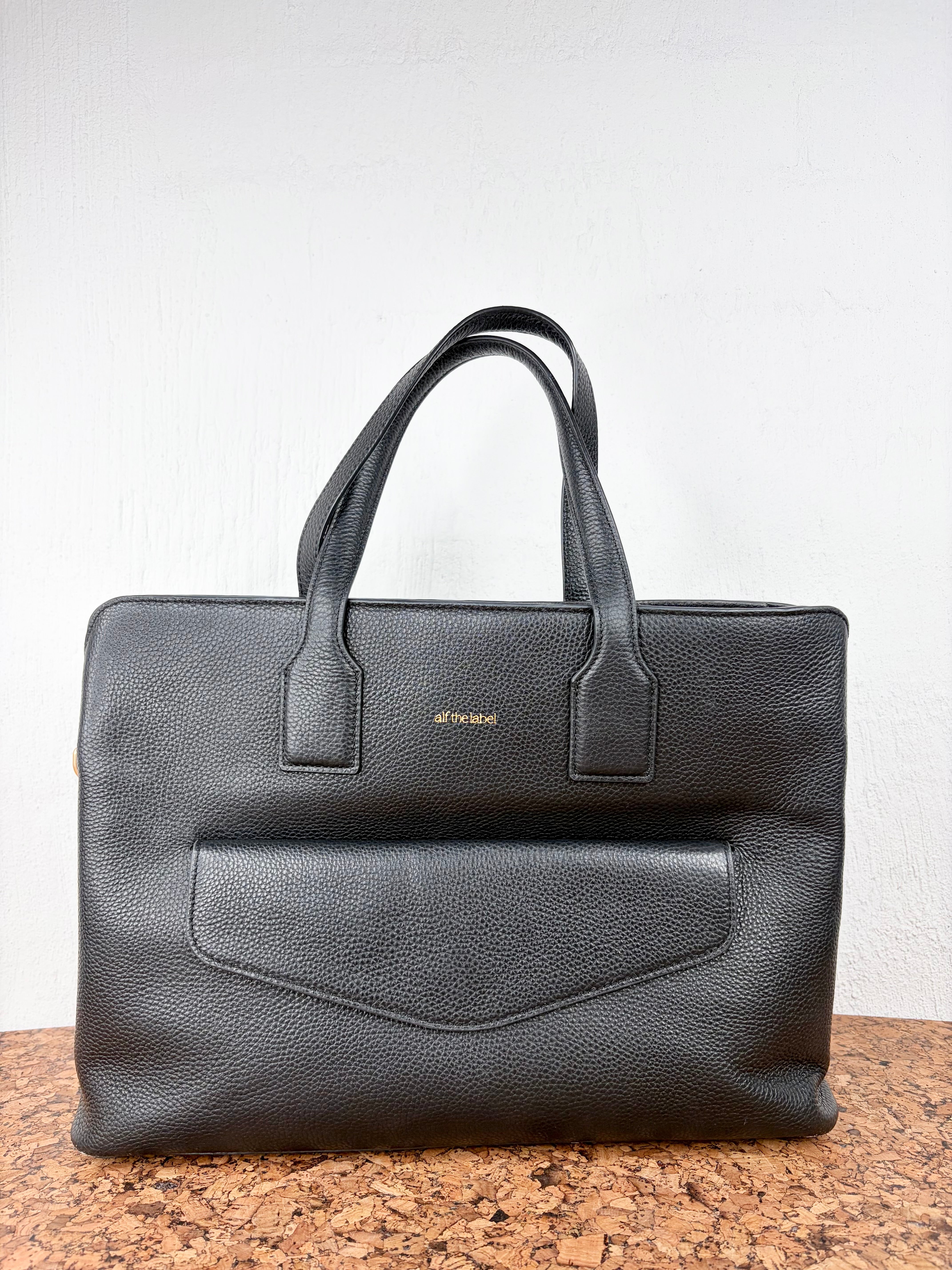 A black pebbled leather tote bag with dual top handles and a front flap pocket, standing on a speckled brown surface against a white wall.
