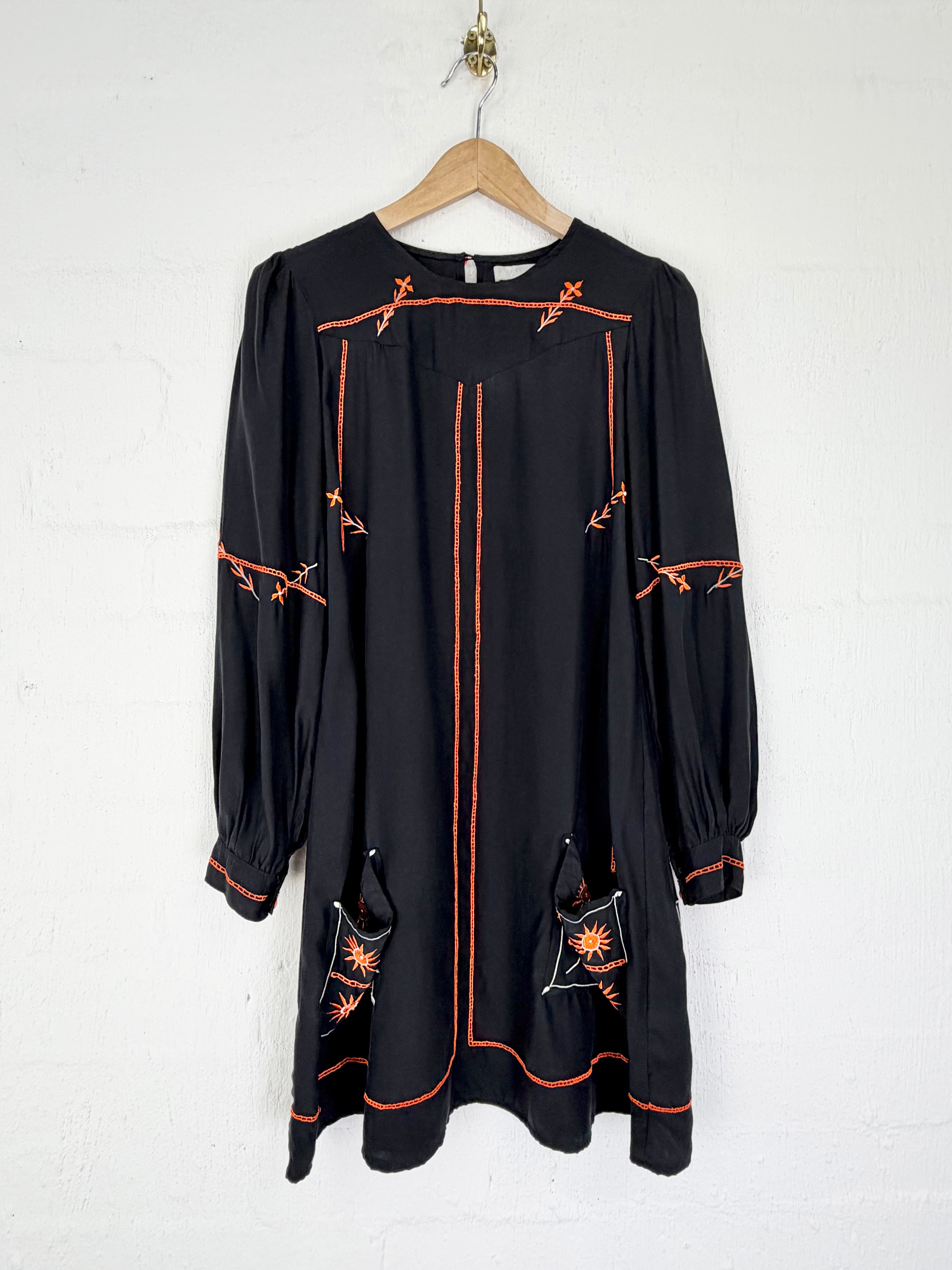 A black long-sleeve dress with orange embroidered spiderweb and star motifs, hanging on a wooden hanger against a white painted brick wall.