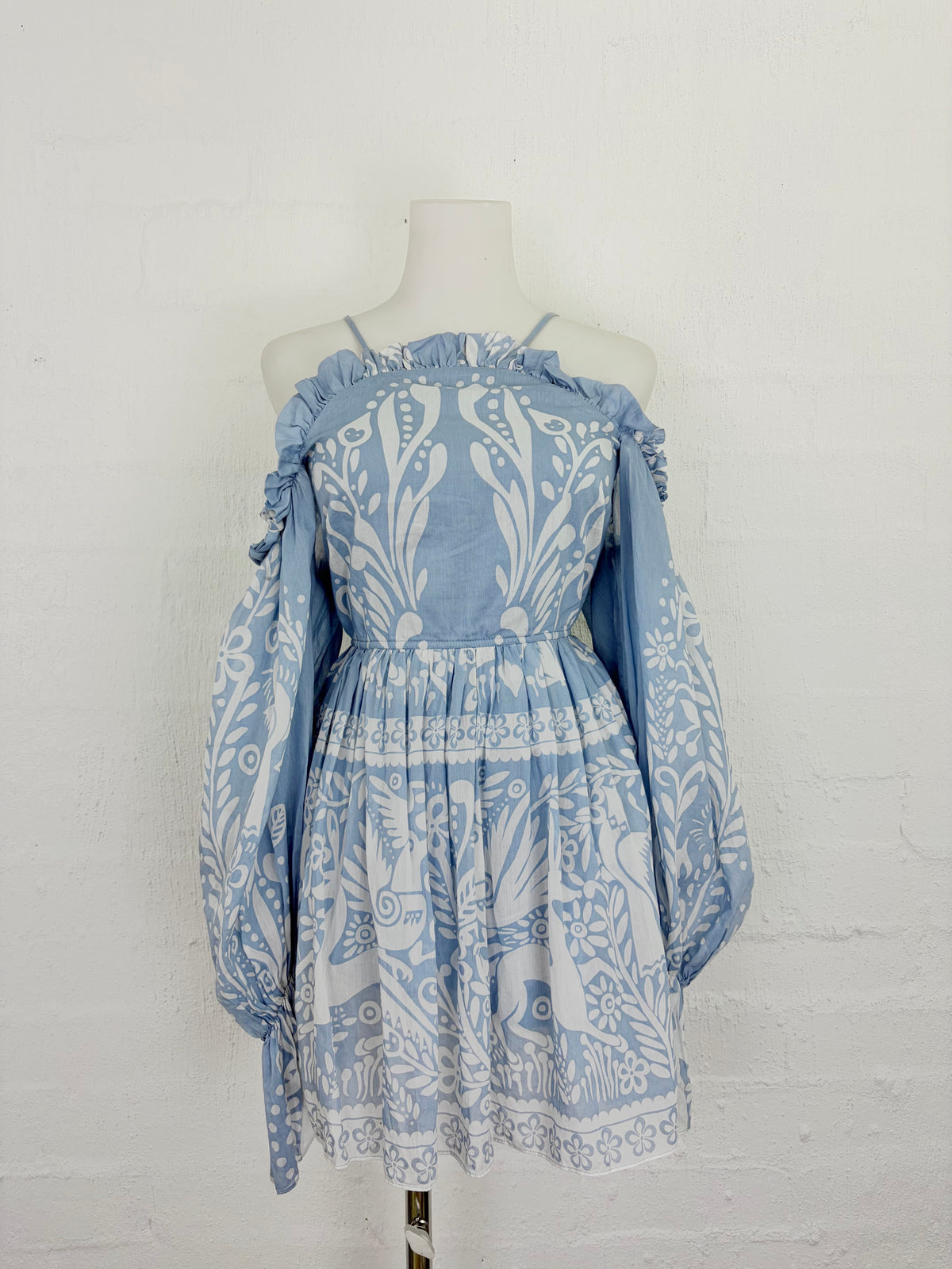 A light blue and white patterned off-the-shoulder dress displayed on a mannequin against a white brick wall.