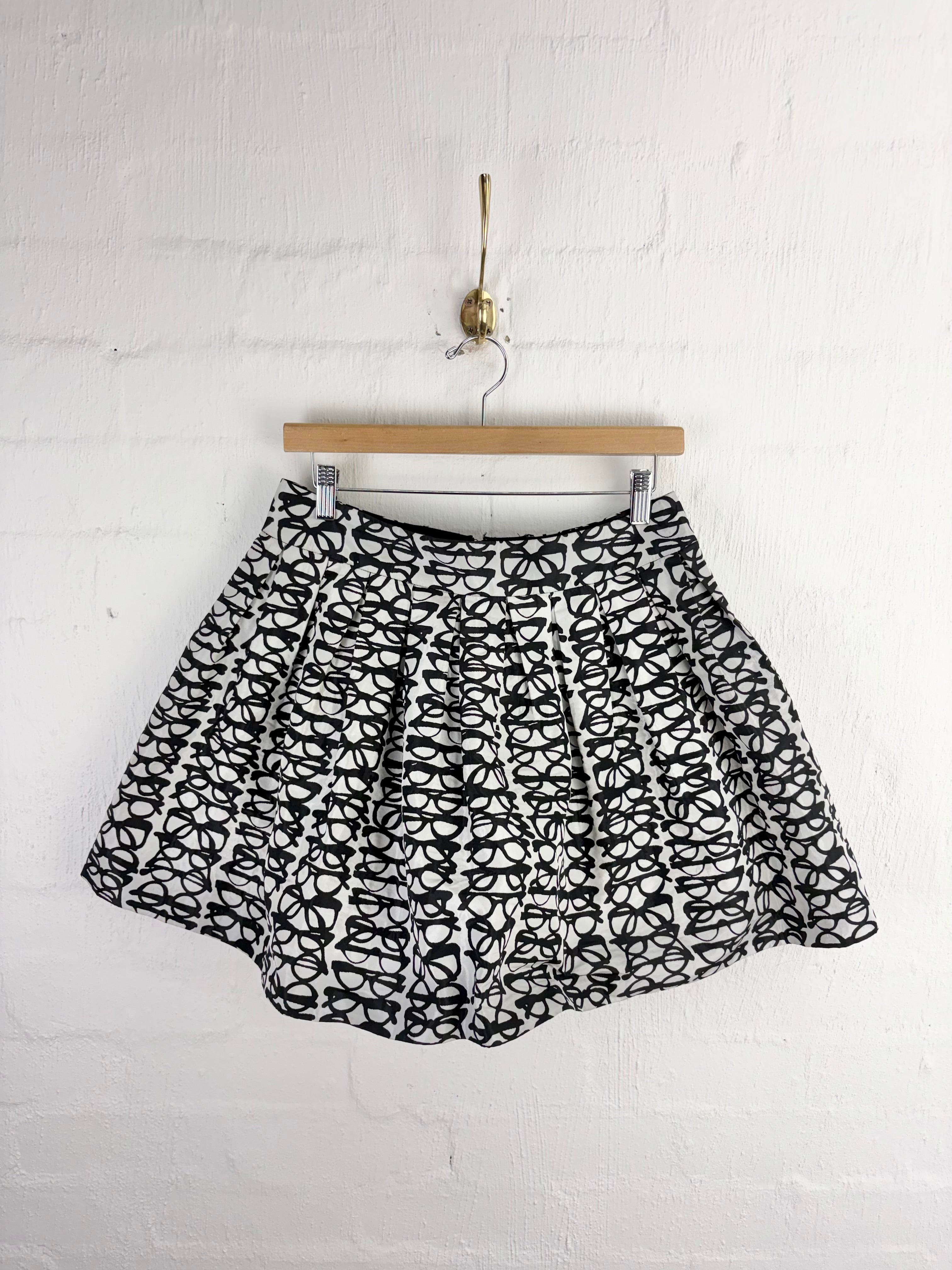 A black and white patterned pleated mini skirt hanging from a wooden hanger against a white painted brick wall.