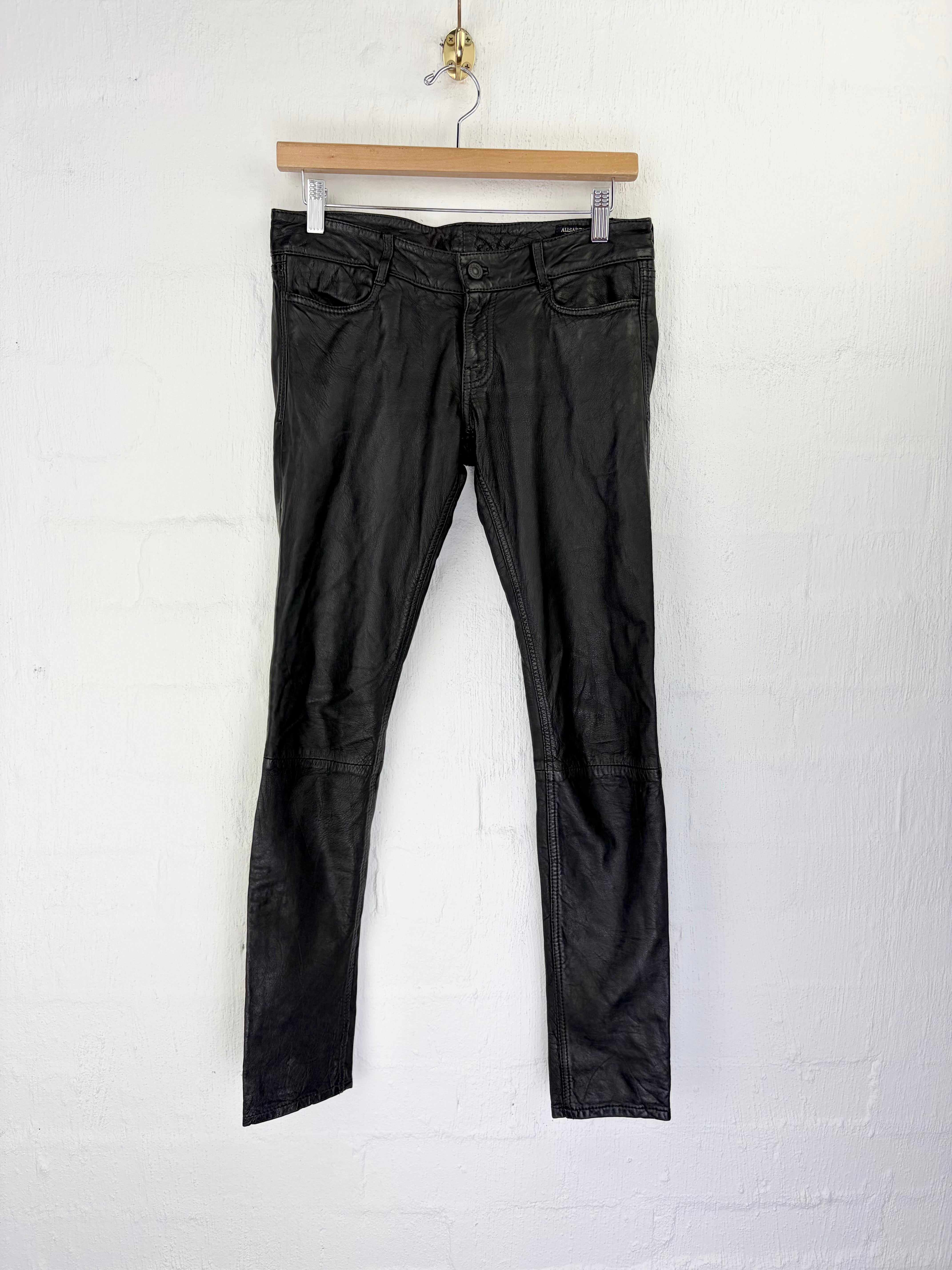 Black leather cropped jeans hanging from a wooden hanger against a white painted brick wall.