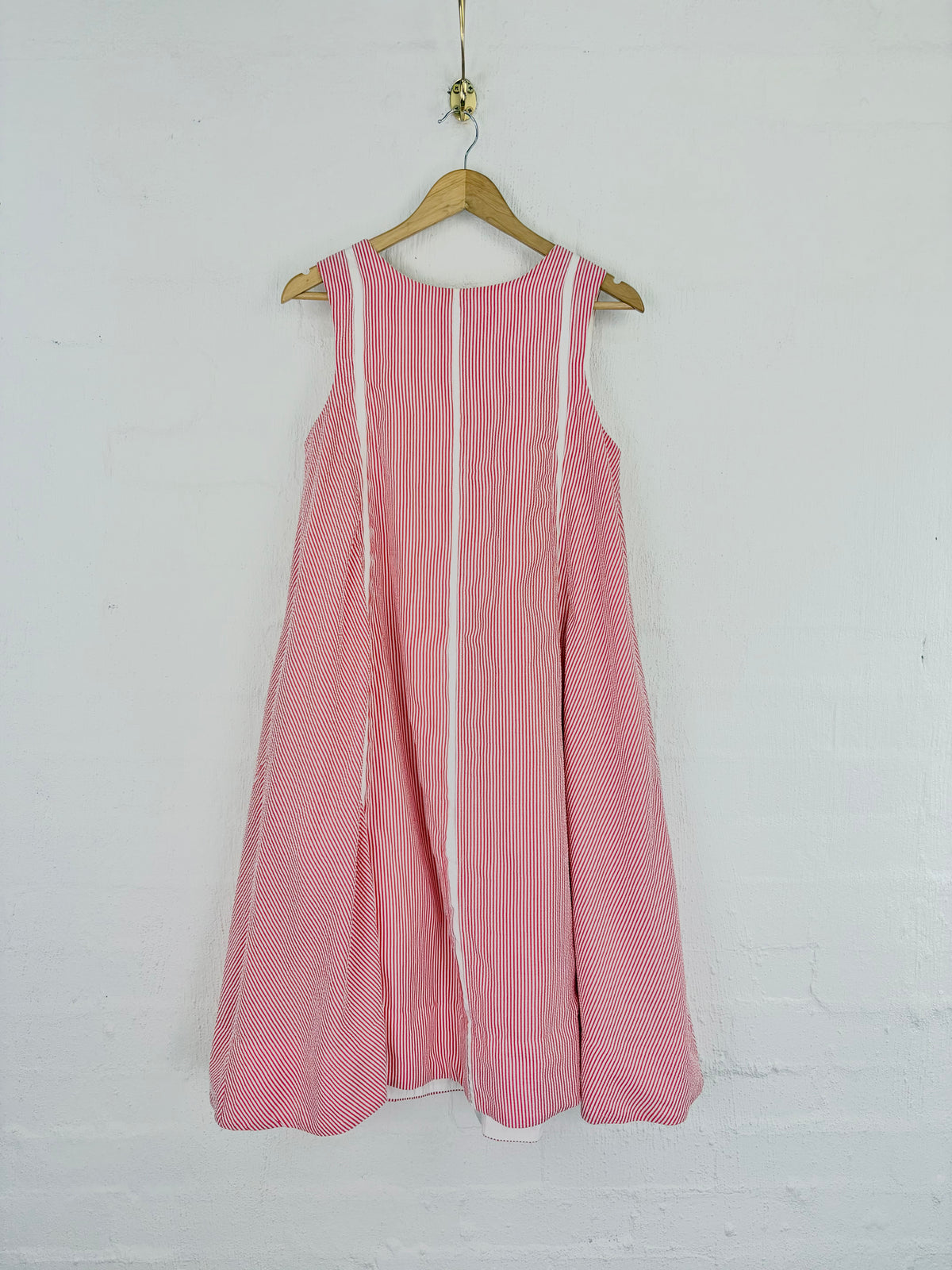 A sleeveless pink dress with white vertical piping details, hanging on a wooden hanger against a white painted brick wall.