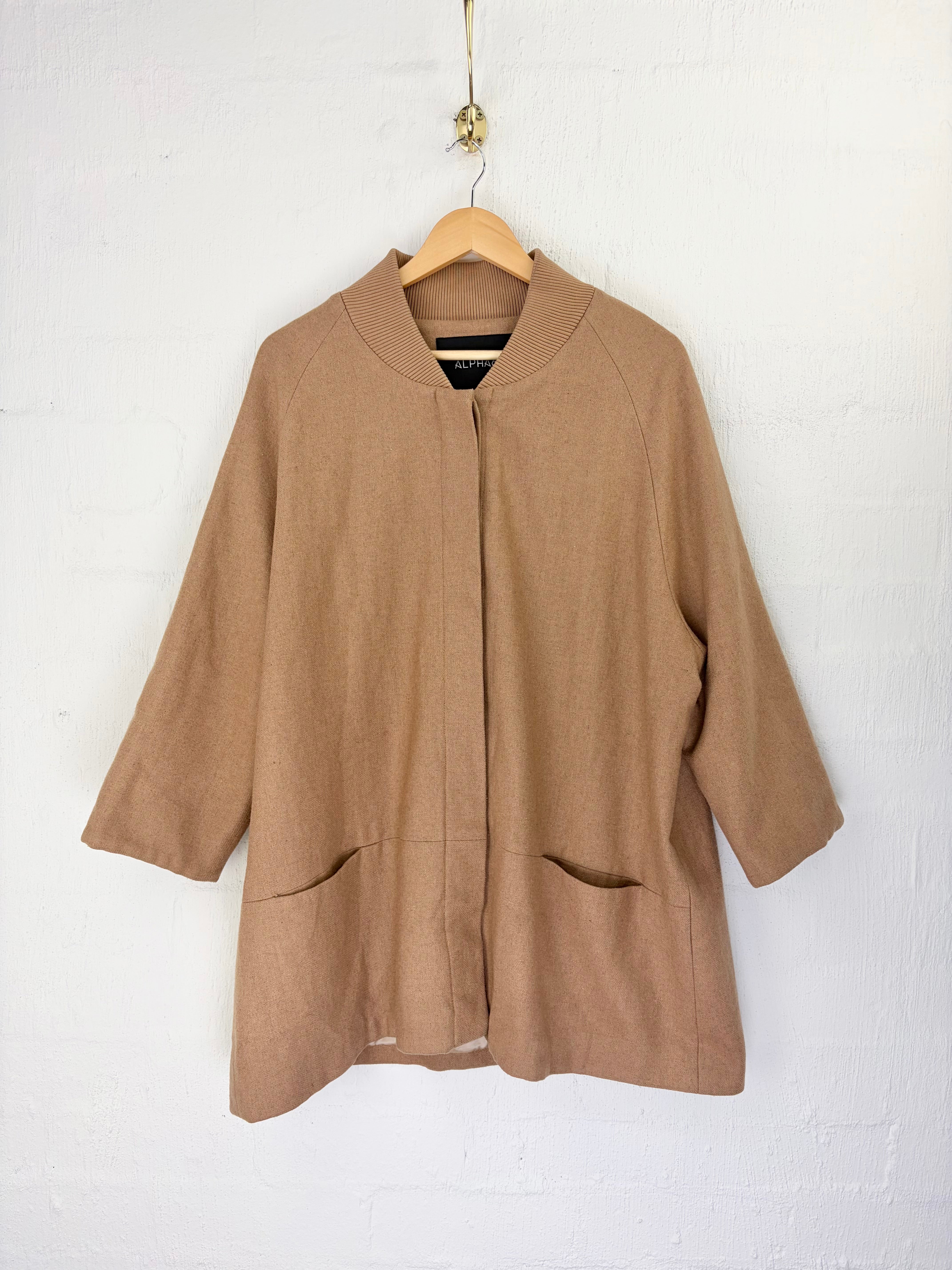 A light brown wool-blend jacket with a mock neck, 3/4 sleeves, and front patch pockets, hanging on a wooden hanger against a white painted brick wall.