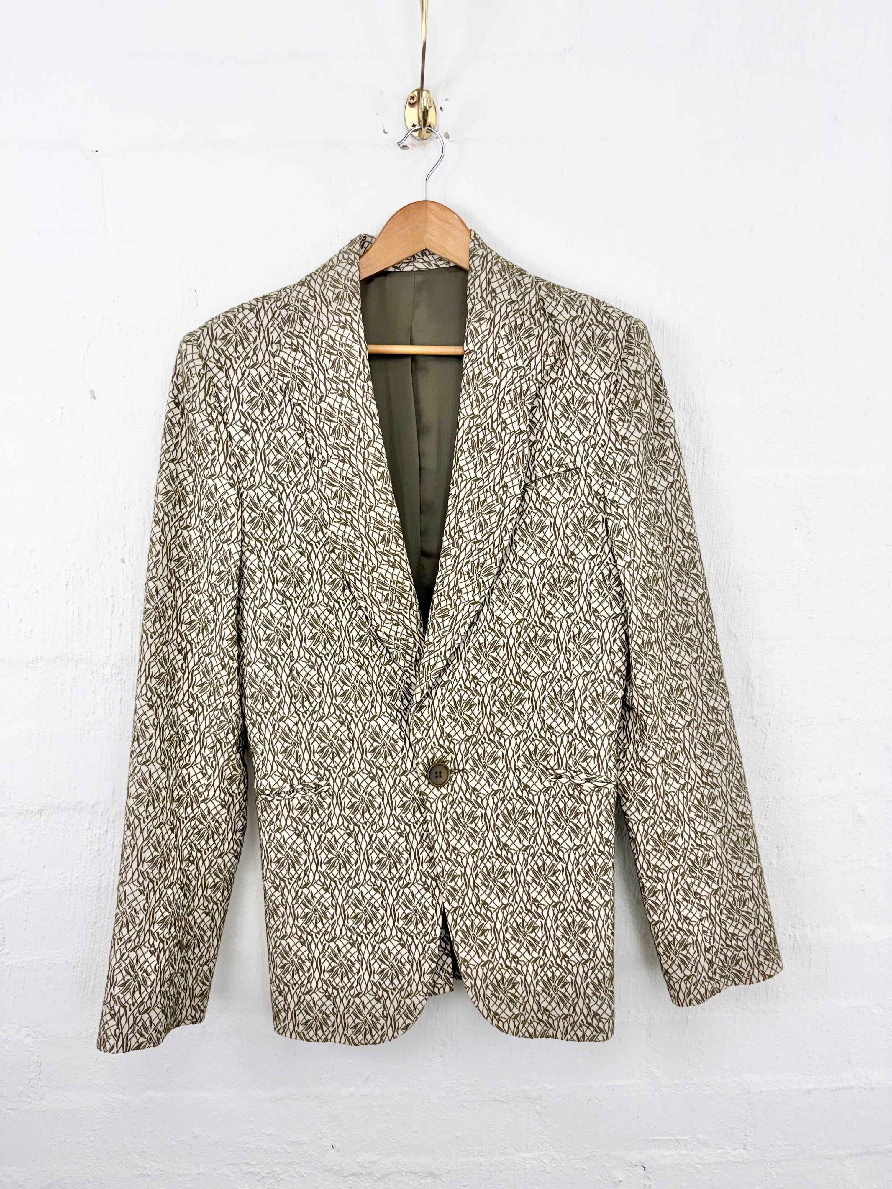 A single-breasted blazer jacket with a textured beige and brown houndstooth pattern, hanging on a wooden hanger against a white painted brick wall.