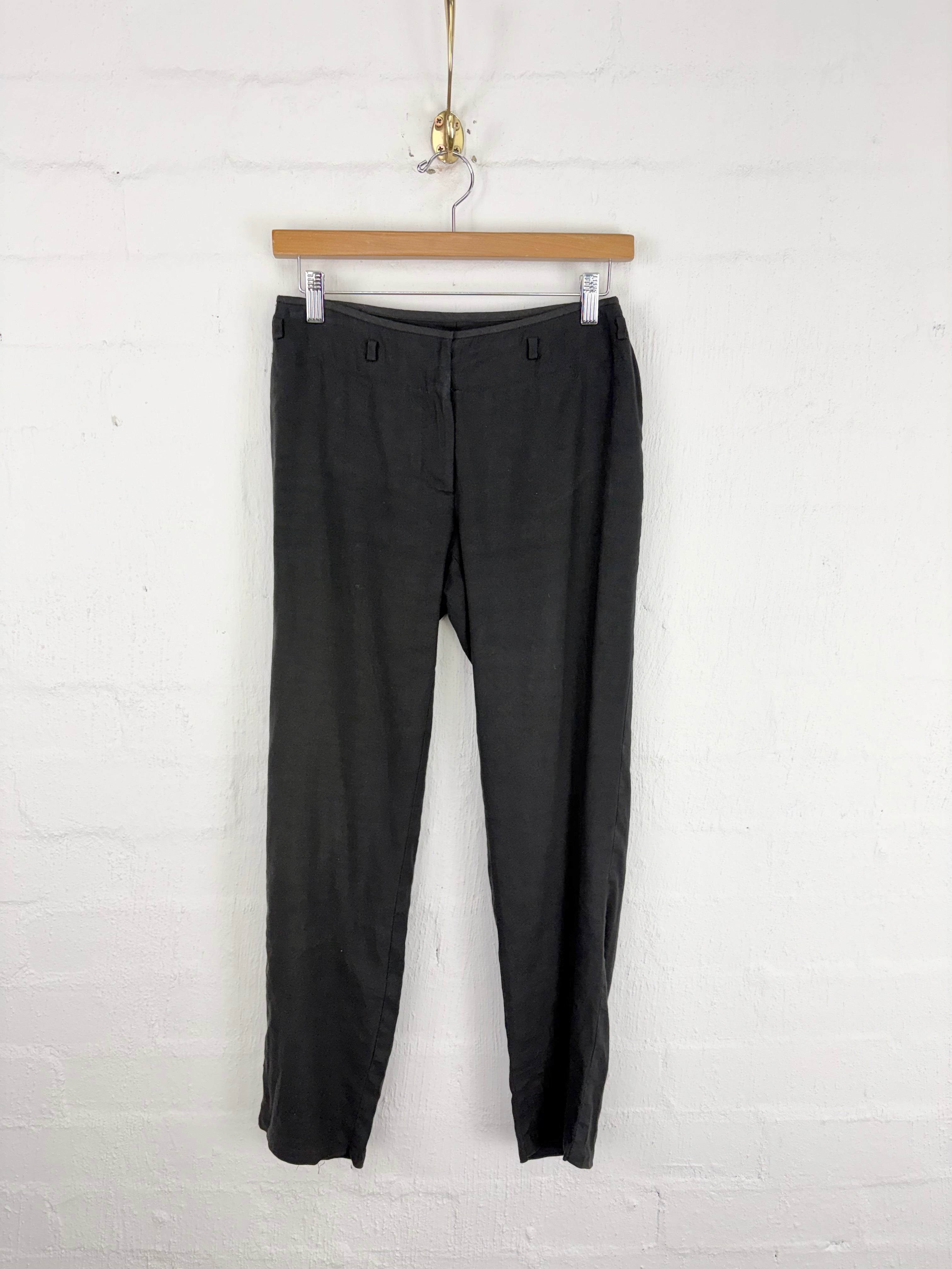 Black slim-fit trousers hanging from a wooden hanger against a white painted brick wall.