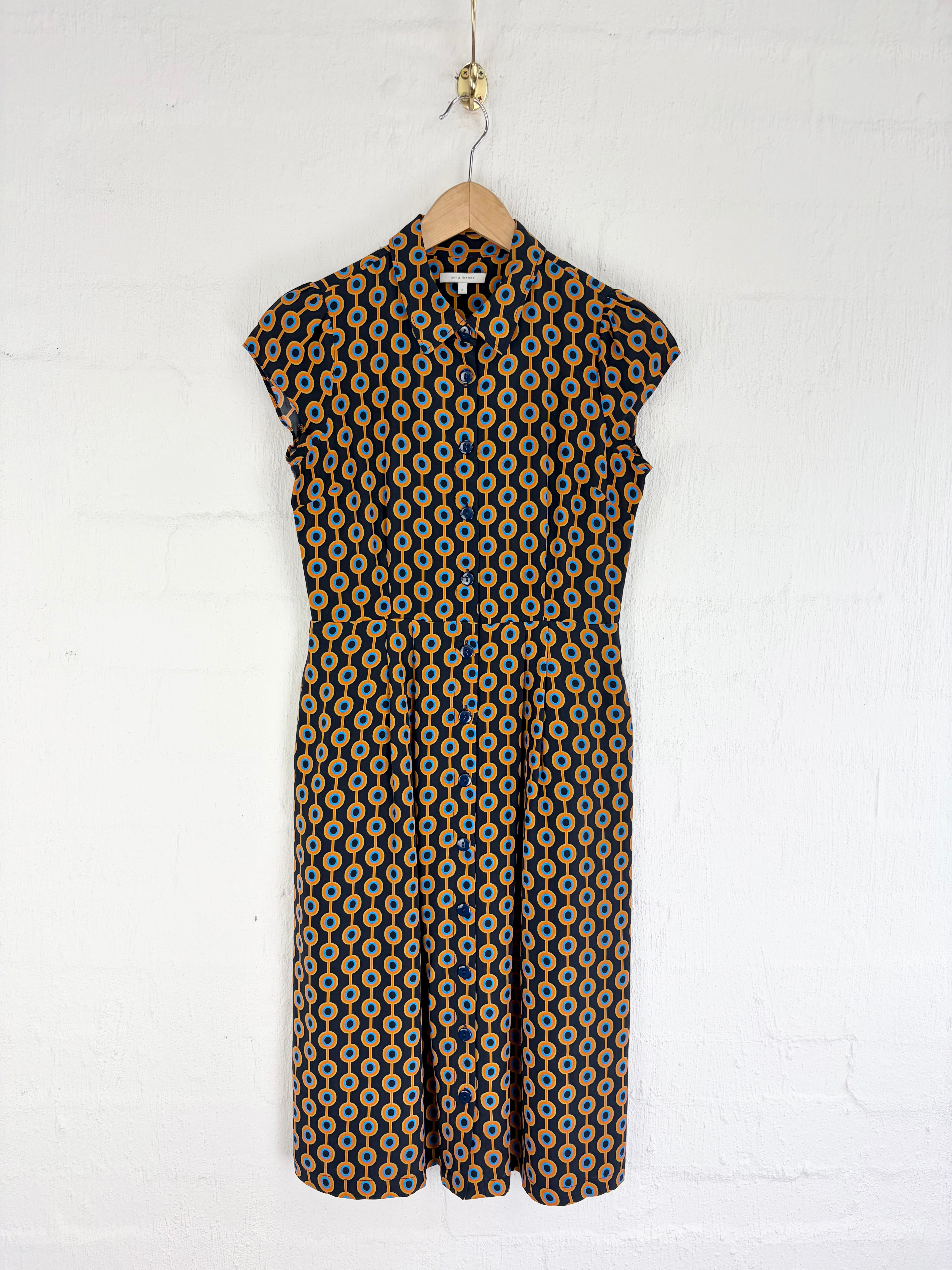 A sleeveless button-front dress with a geometric circular pattern in black, mustard yellow, and white, hanging on a wooden hanger against a white painted brick wall.