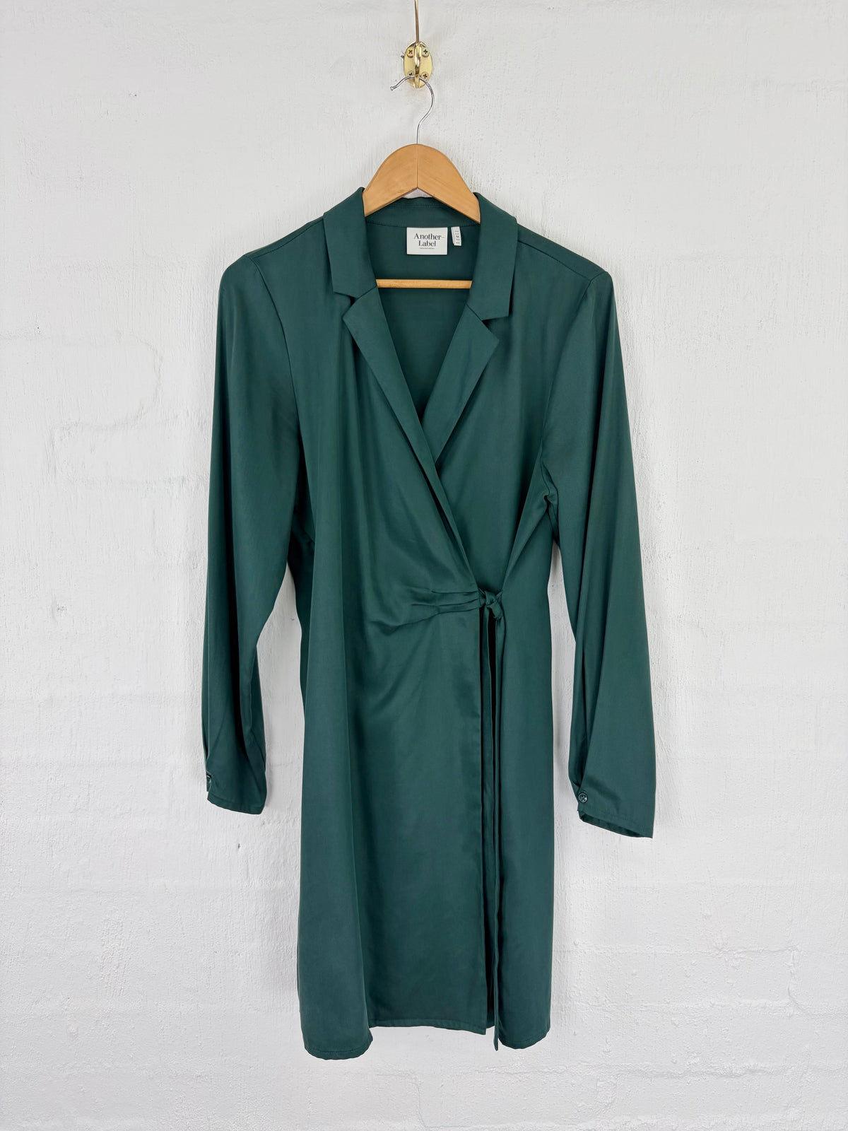 A dark green wrap-style long-sleeve dress with a tie waist, hanging on a wooden hanger against a white painted brick wall.