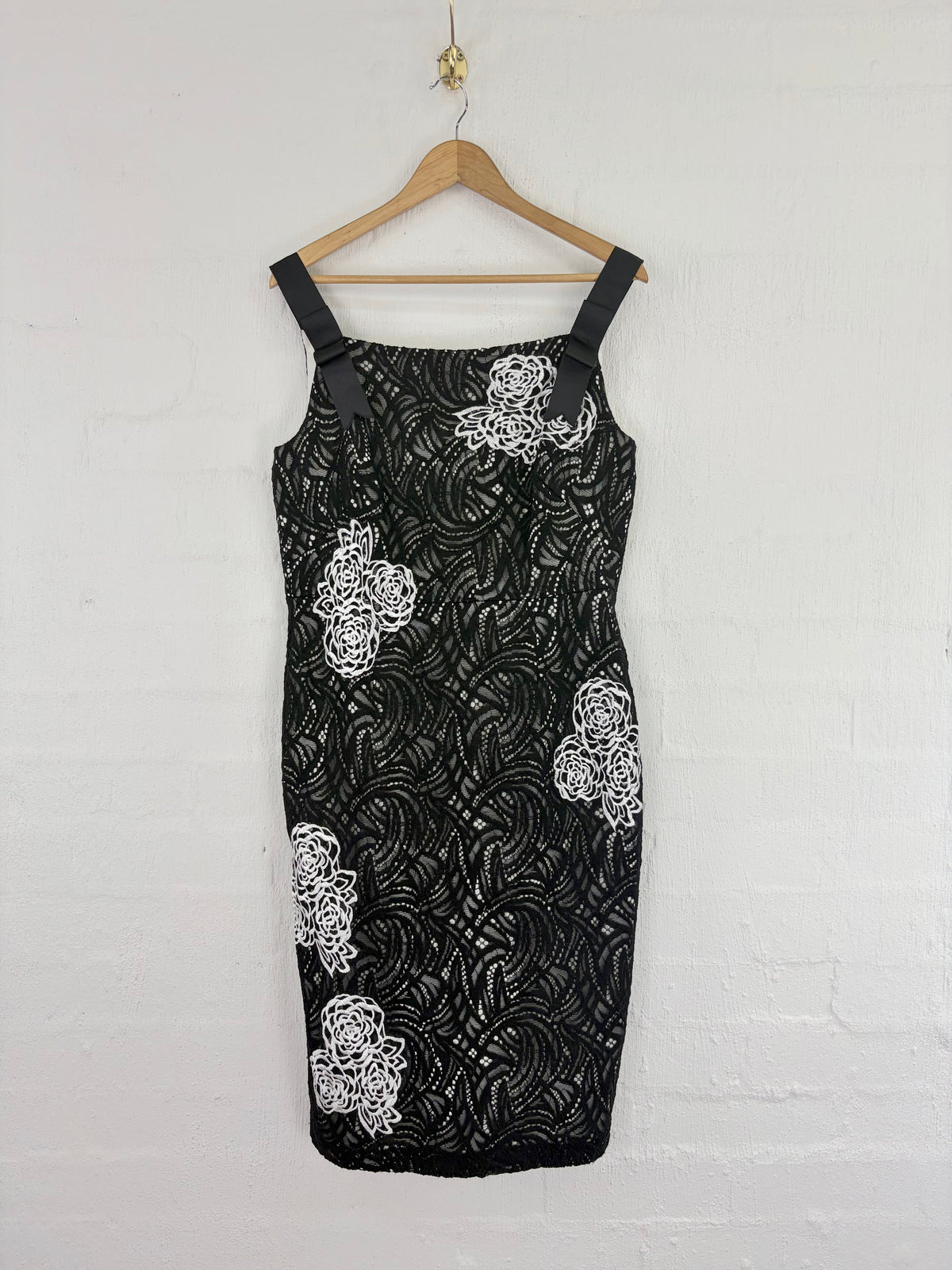 A sleeveless black lace dress with white floral applique details, hanging on a wooden hanger against a white painted brick wall.