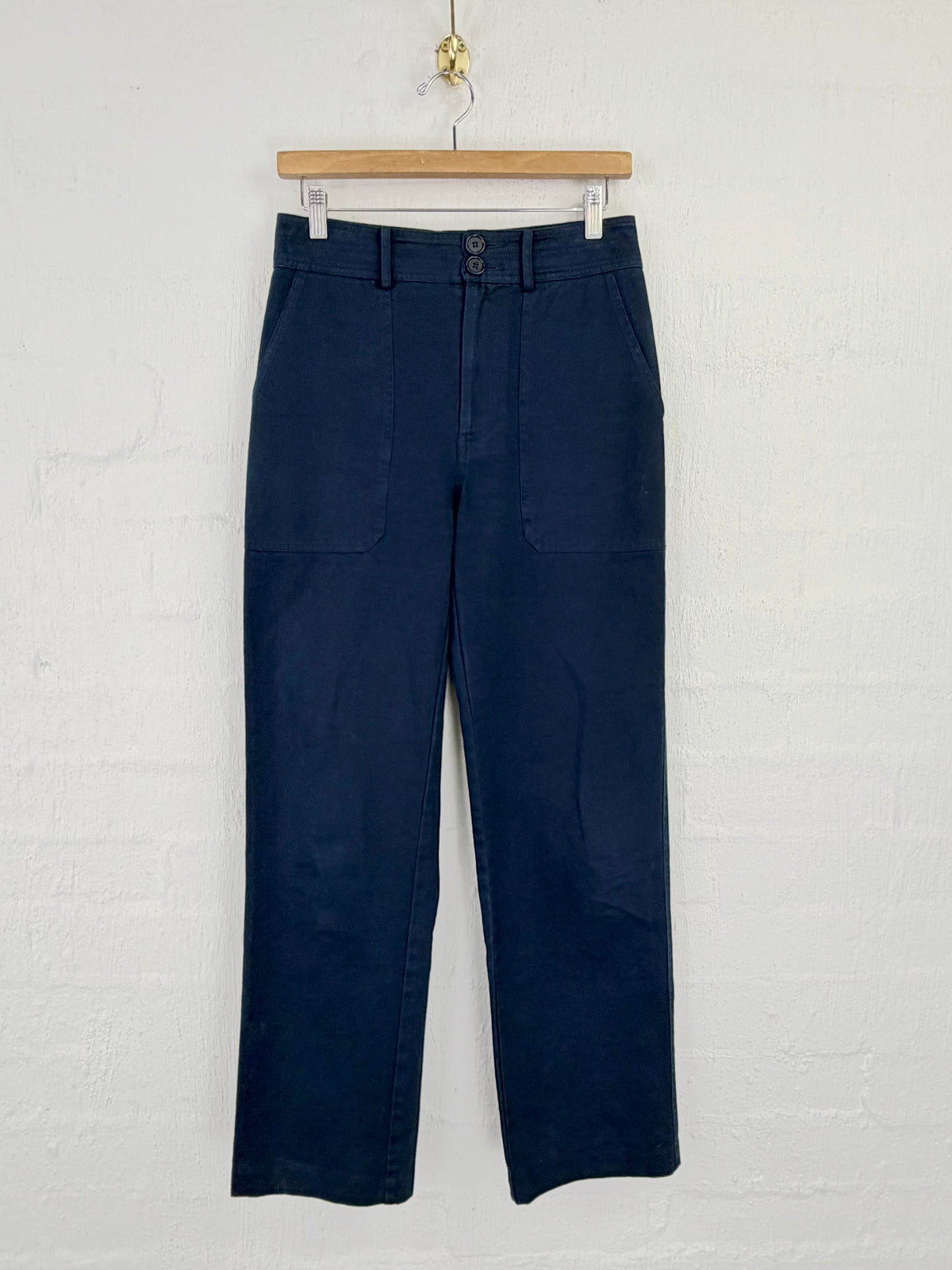 A pair of dark navy blue cropped pants hanging from a wooden hanger against a white painted brick wall.