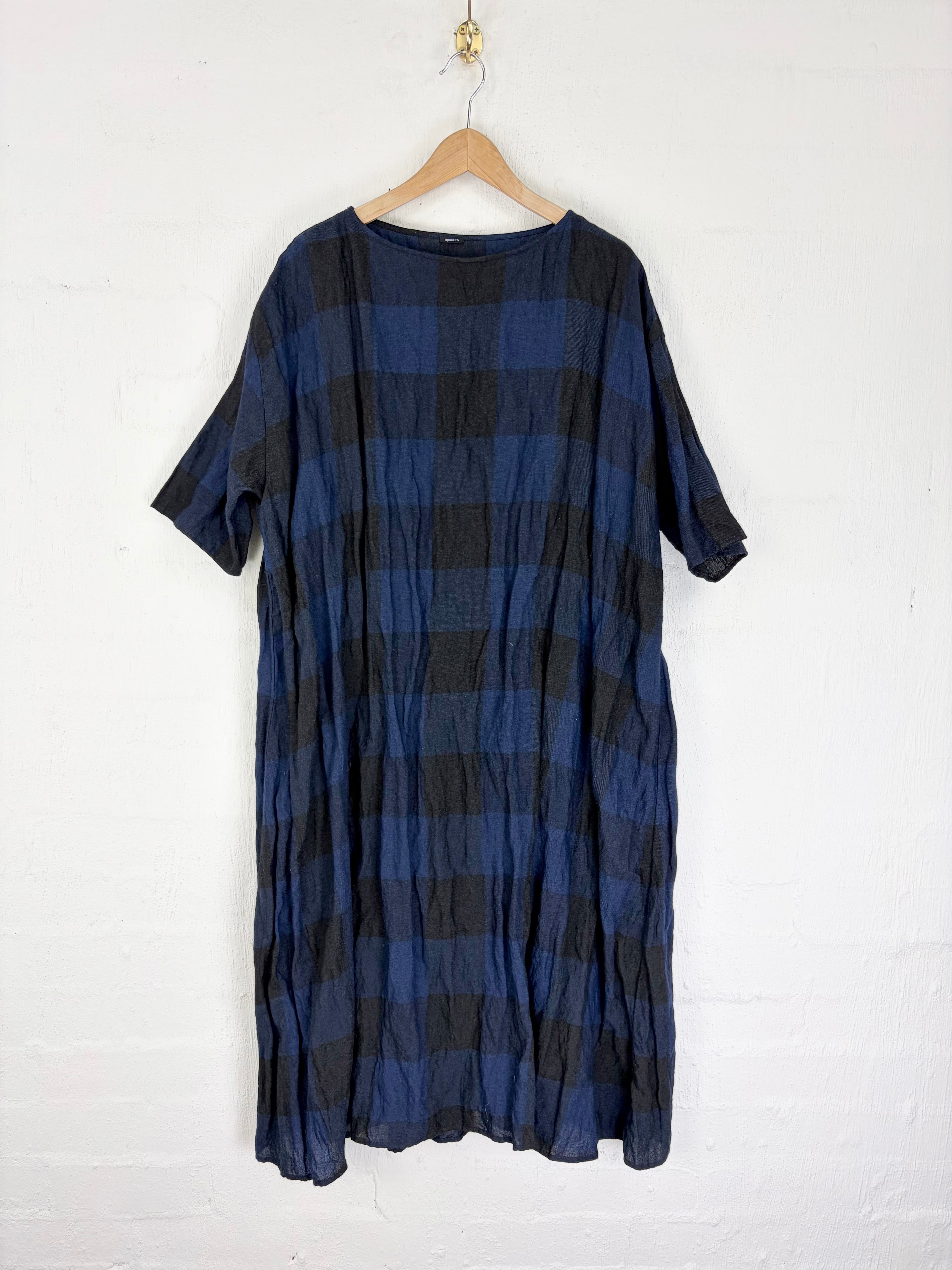 A loose short-sleeve dress with a blue and black plaid pattern, hanging on a wooden hanger against a white painted brick wall.