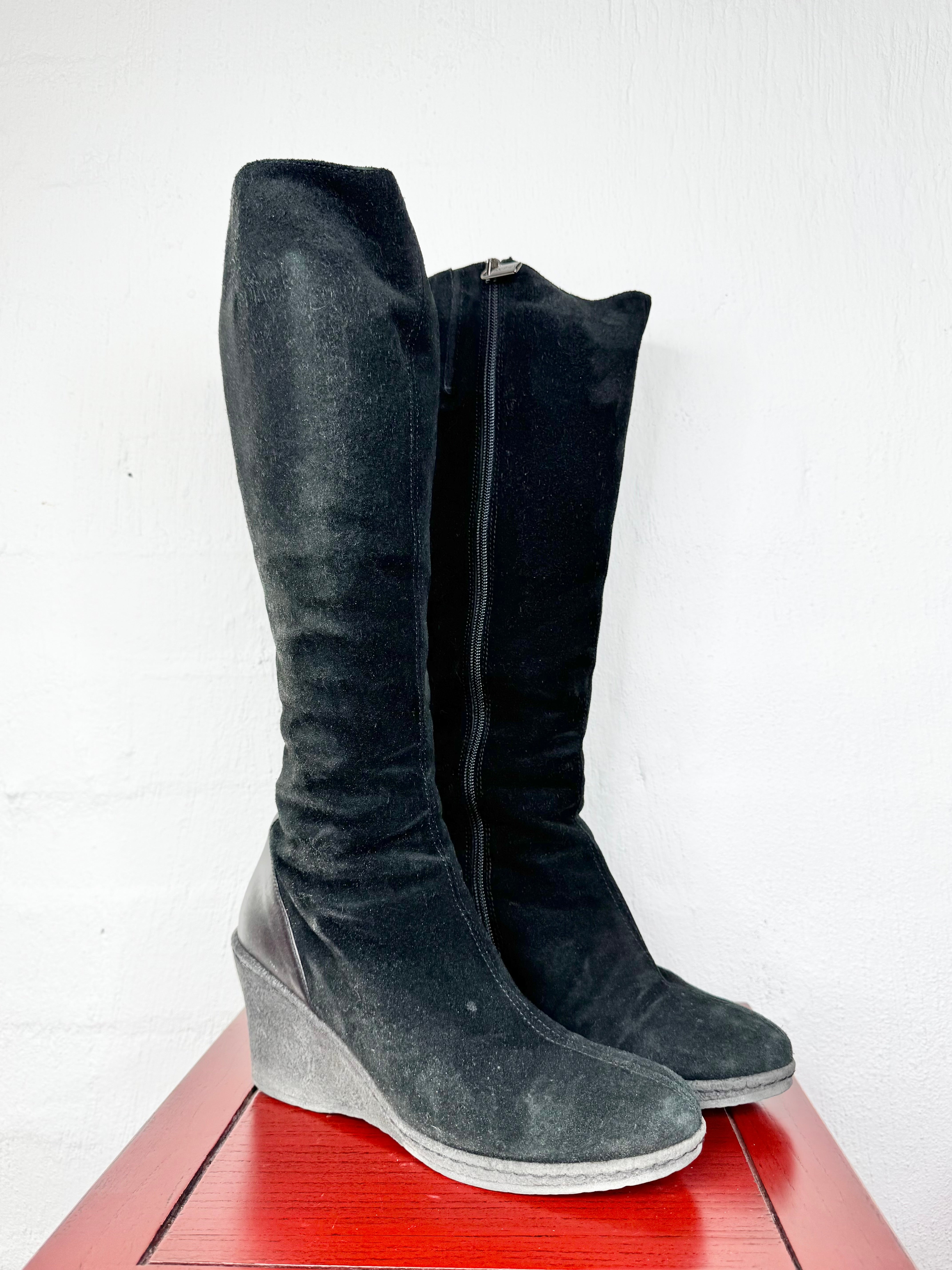 A pair of tall black suede knee-high boots with wedge heels, standing on a red wooden surface against a white wall.