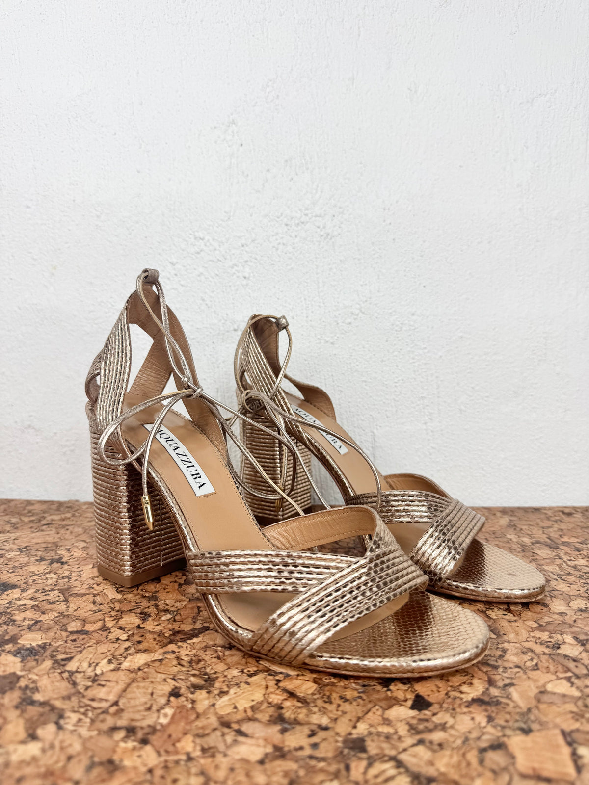 A pair of metallic gold strappy high-heeled sandals with lace-up ankle straps, displayed on a cork surface against a white wall.