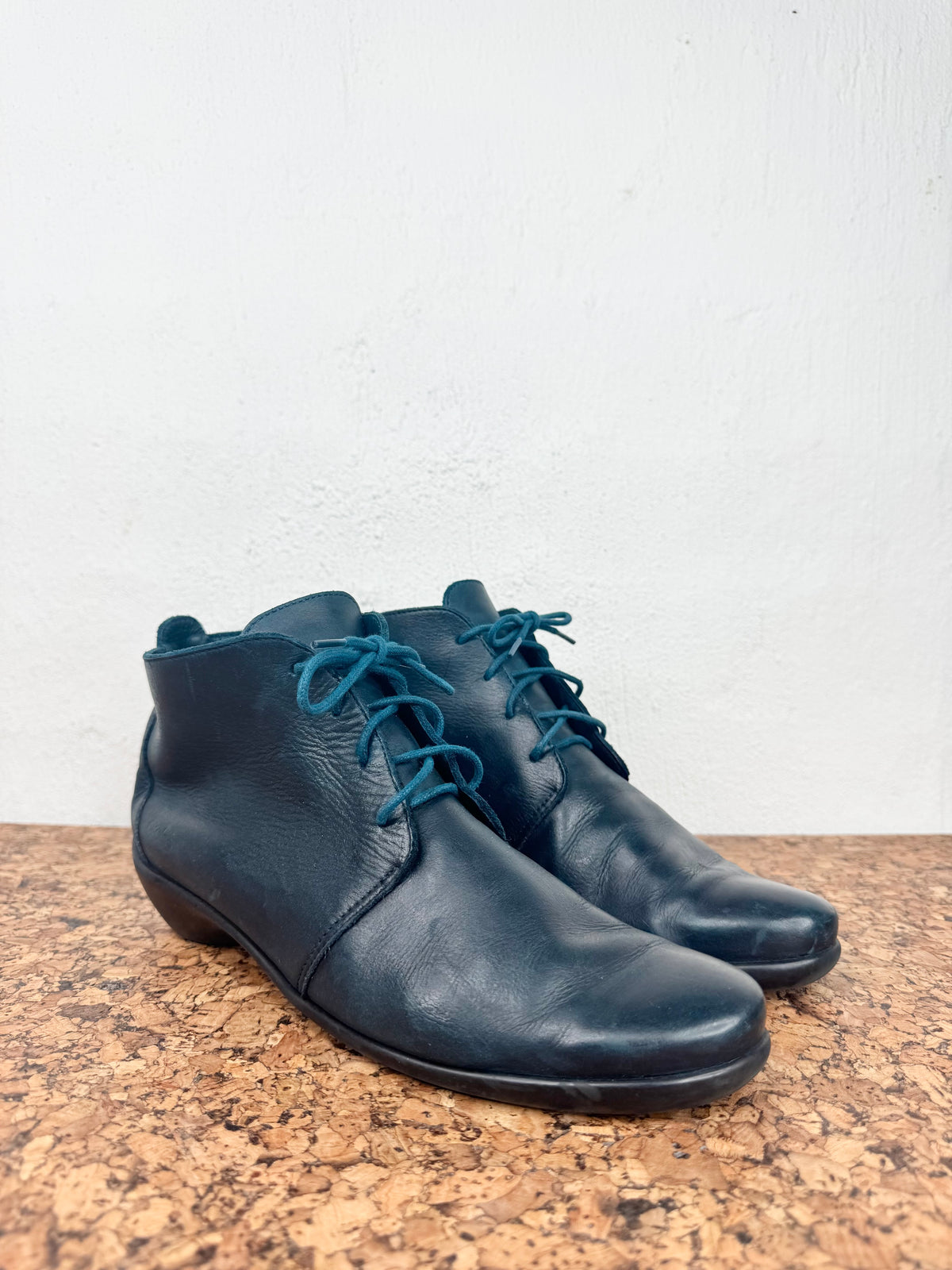 A pair of dark navy blue leather ankle boots with teal-blue laces, resting on a speckled brown countertop against a white wall.