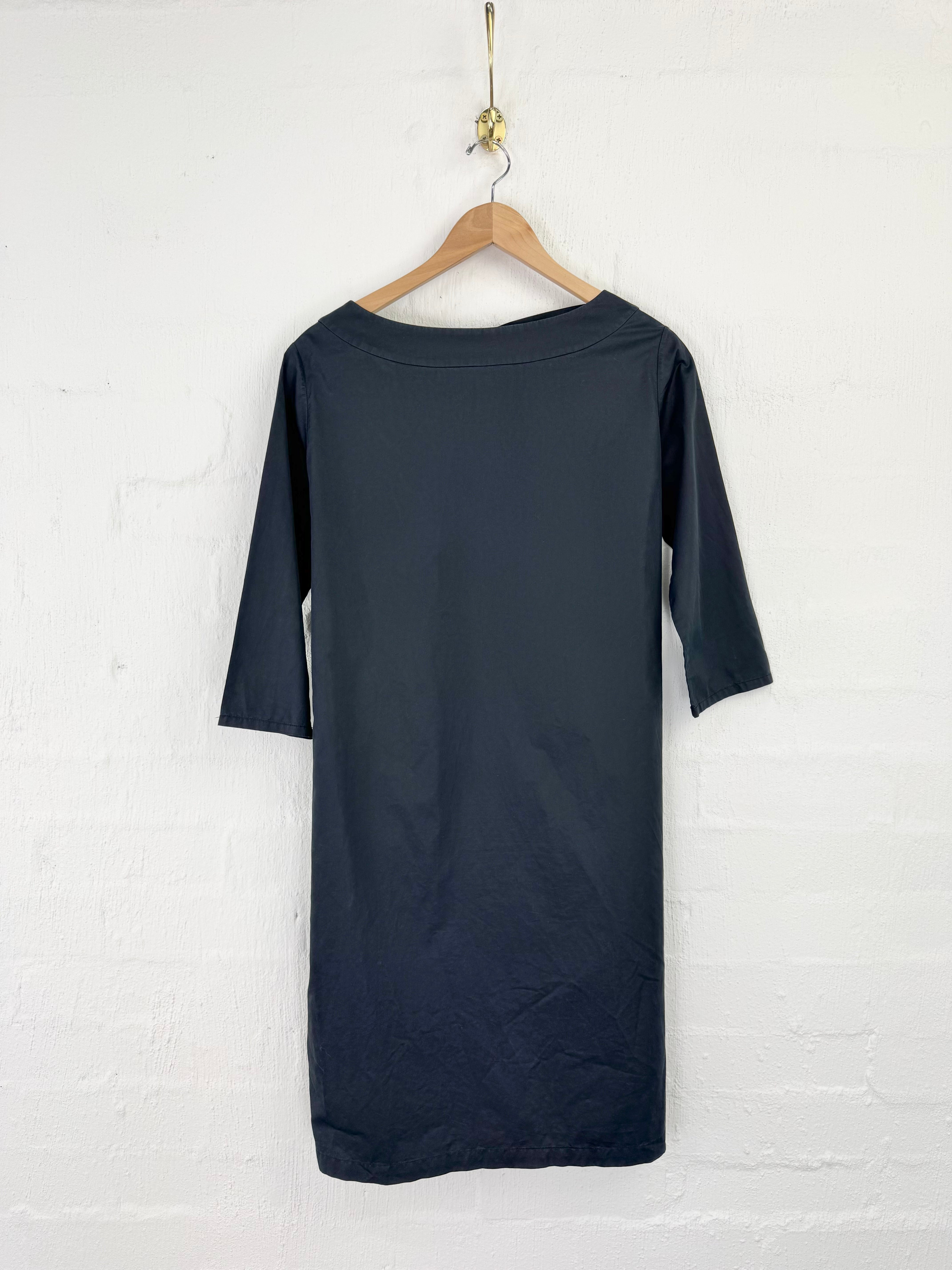 A dark navy blue dress with three-quarter sleeves hanging on a wooden hanger against a white brick wall.