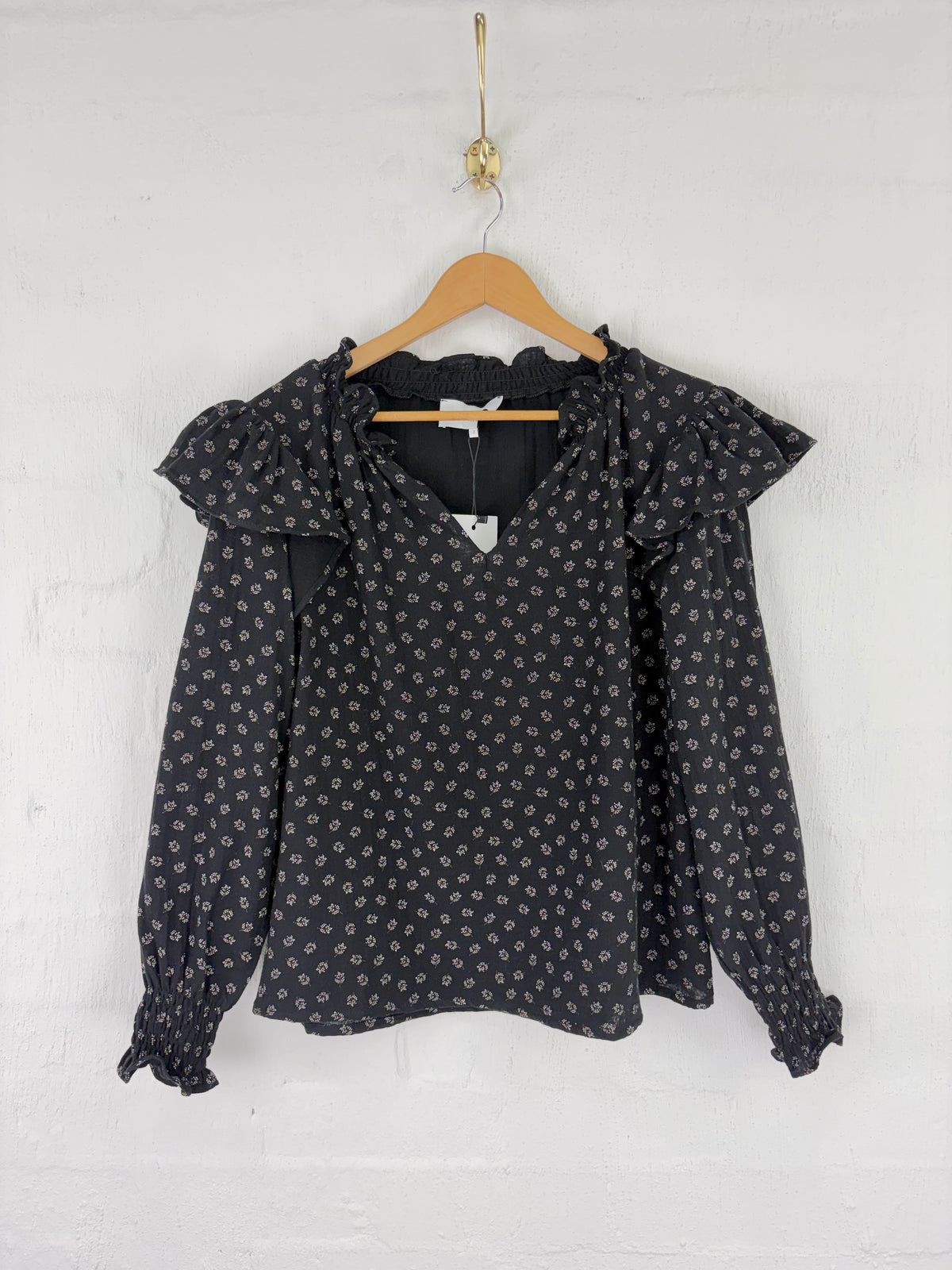 A black long-sleeve blouse with a small white floral print, featuring ruffled shoulders and a V-neckline, hanging on a wooden hanger against a white painted brick wall.