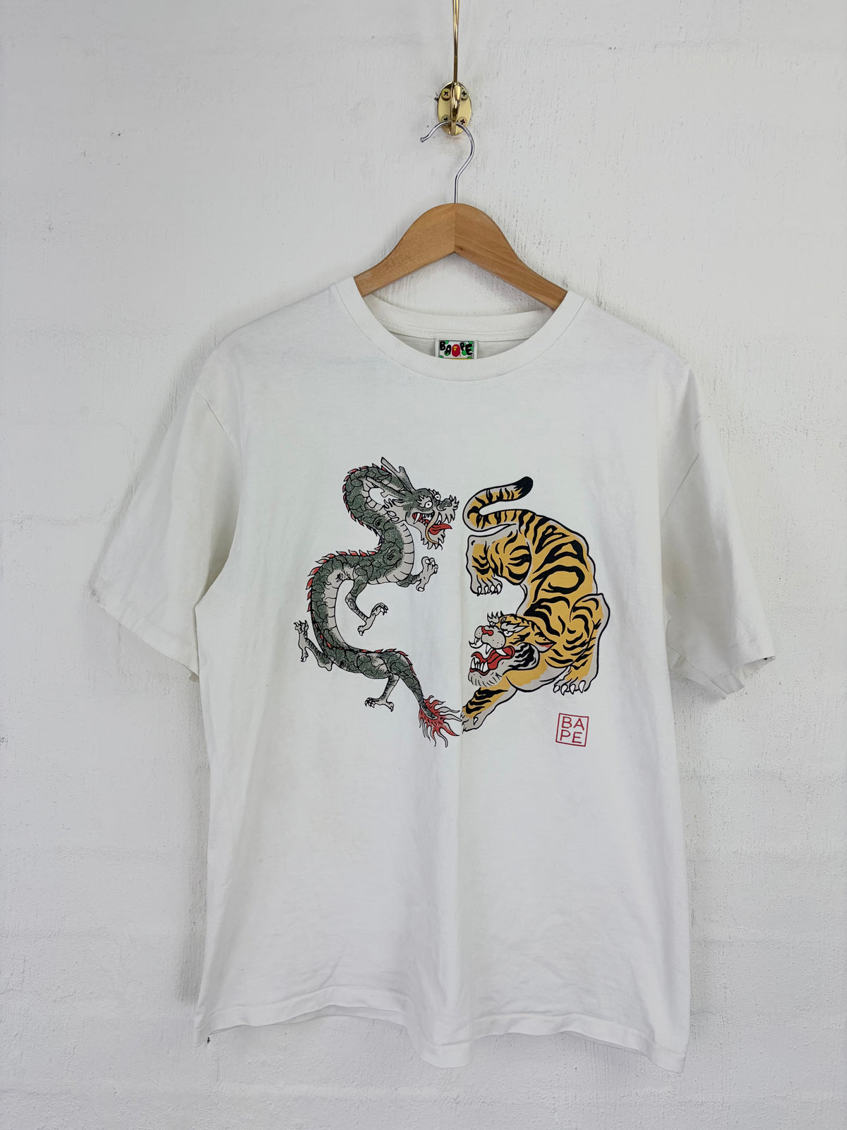 A white short-sleeve t-shirt hanging on a wooden hanger against a white brick wall. The front of the shirt features a graphic print of a black and gray dragon facing off against a yellow and black tiger, with a small red seal at the bottom right of the graphic.