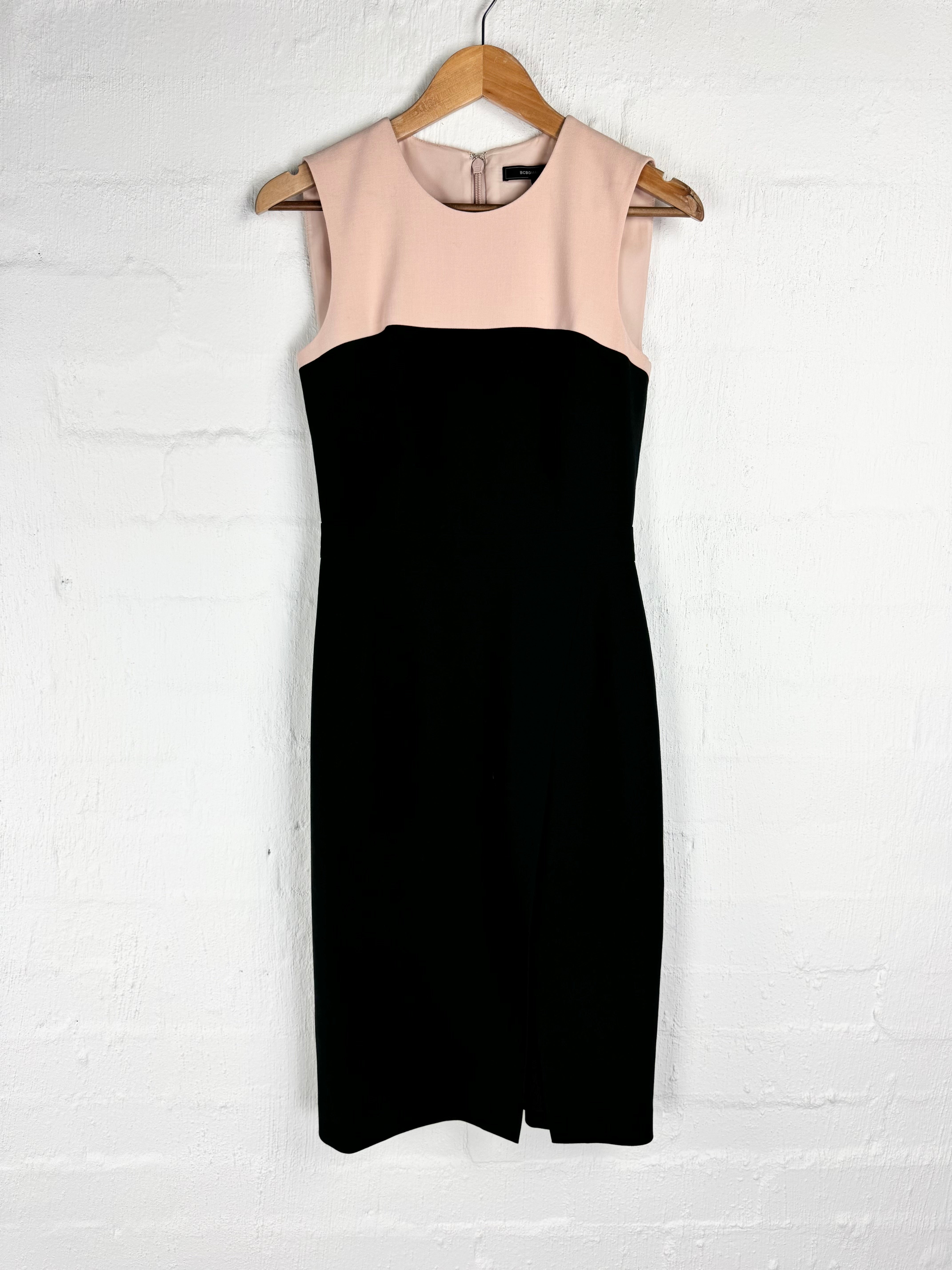 A color-block sleeveless dress hanging on a wooden hanger against a white painted brick wall. The bodice is a soft blush pink, and the skirt is solid black.