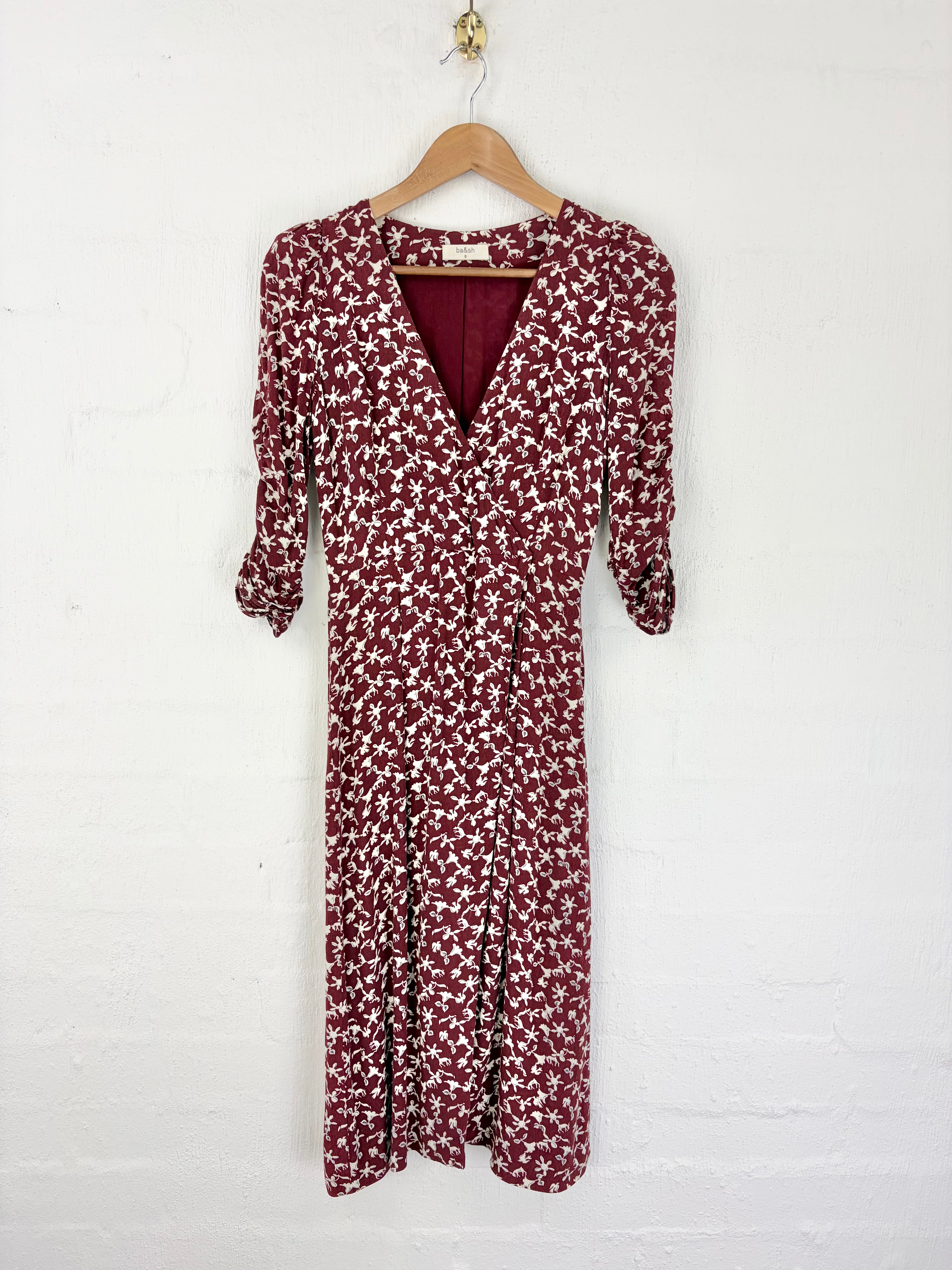 A long-sleeve wrap dress with a dense small floral print in burgundy and white, hanging on a wooden hanger against a white painted brick wall.
