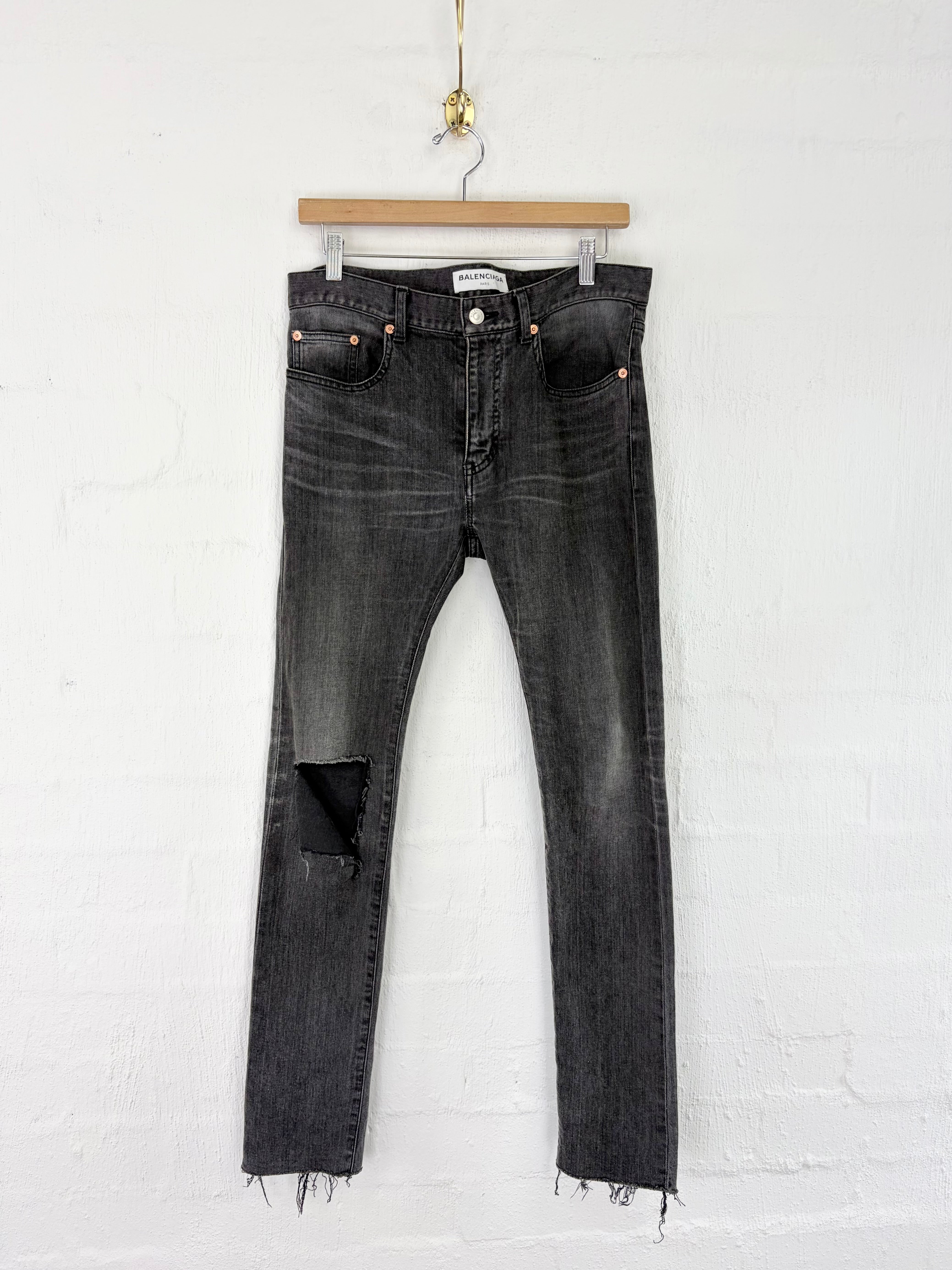 A pair of dark gray washed denim jeans with a ripped knee detail and raw frayed hems, hanging from a wooden hanger against a white brick wall.
