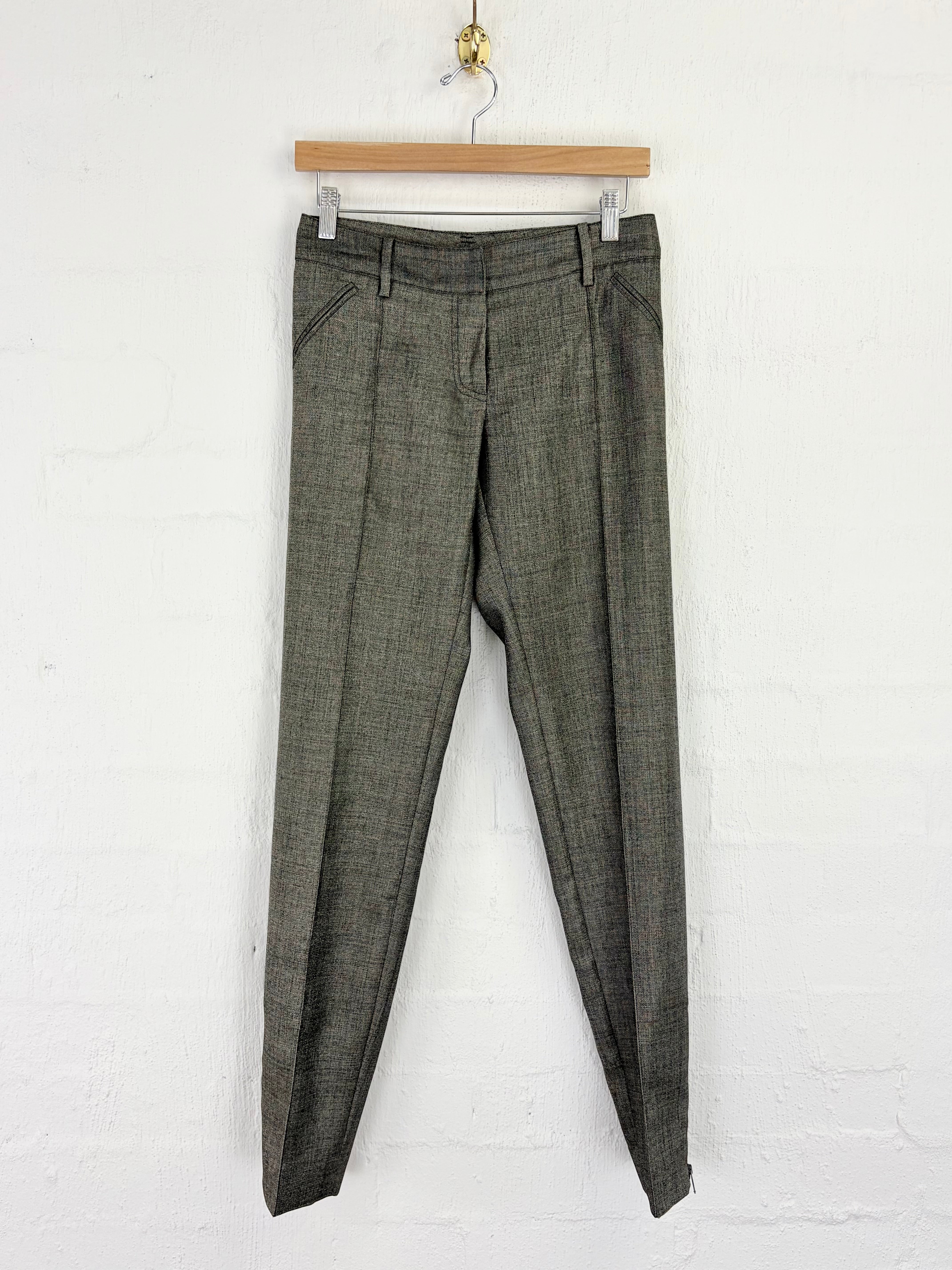 A pair of olive green plaid trousers hanging from a wooden hanger against a white painted brick wall.