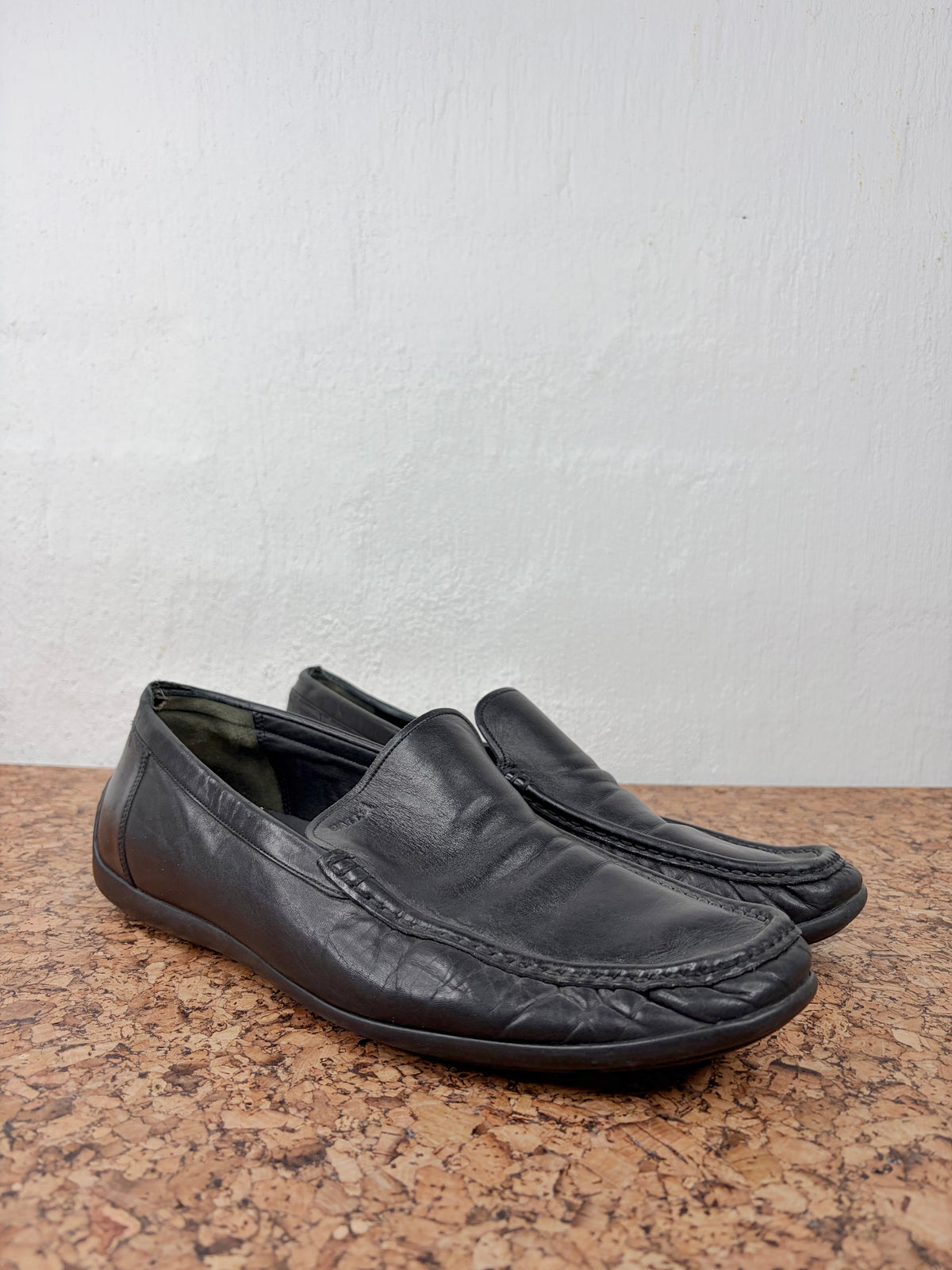 A pair of black leather driving loafers with a moccasin-style toe and decorative stitching, resting on a brown speckled stone surface against a white wall.